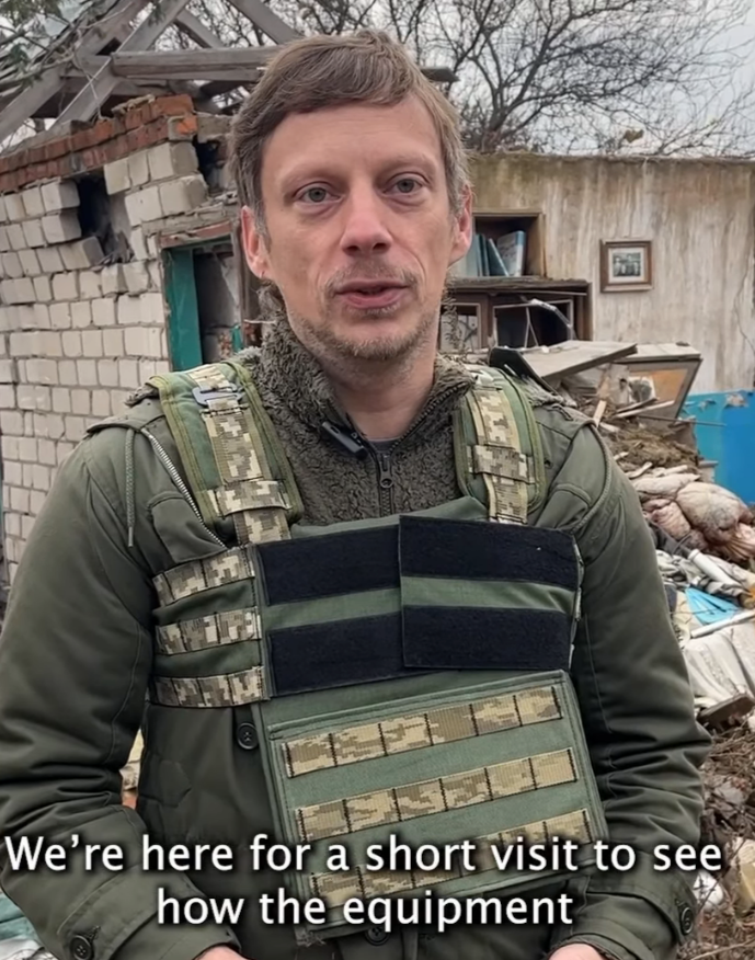 Man in militaire uitrusting met body armor in een buitenomgeving met afval en beschadigde gebouwen.