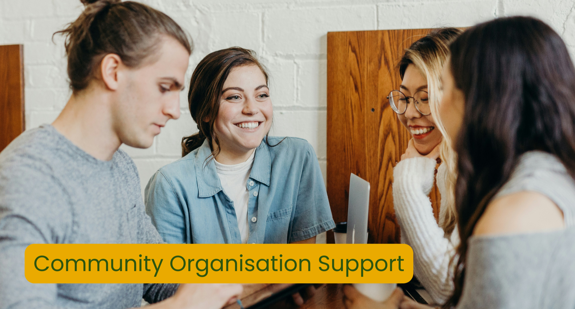 Four young adults sitting around a table, smiling and engaging in conversation, with a sign that reads "Community Organisation Support" in front of them.