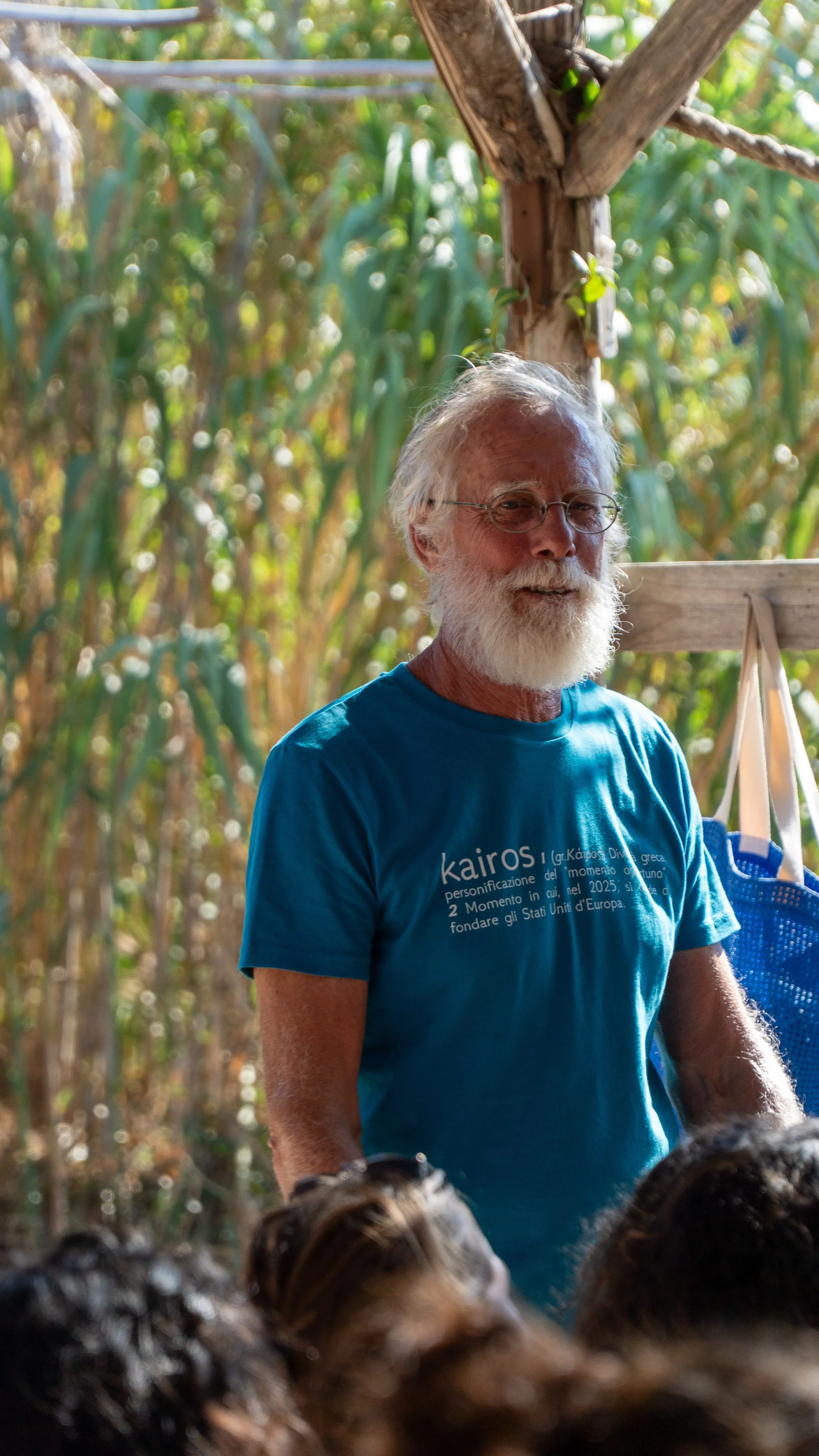Uomo con barba e occhiali, che indossa una maglietta blu con scritto 'kairos', in un ambiente naturale verde con alberi.