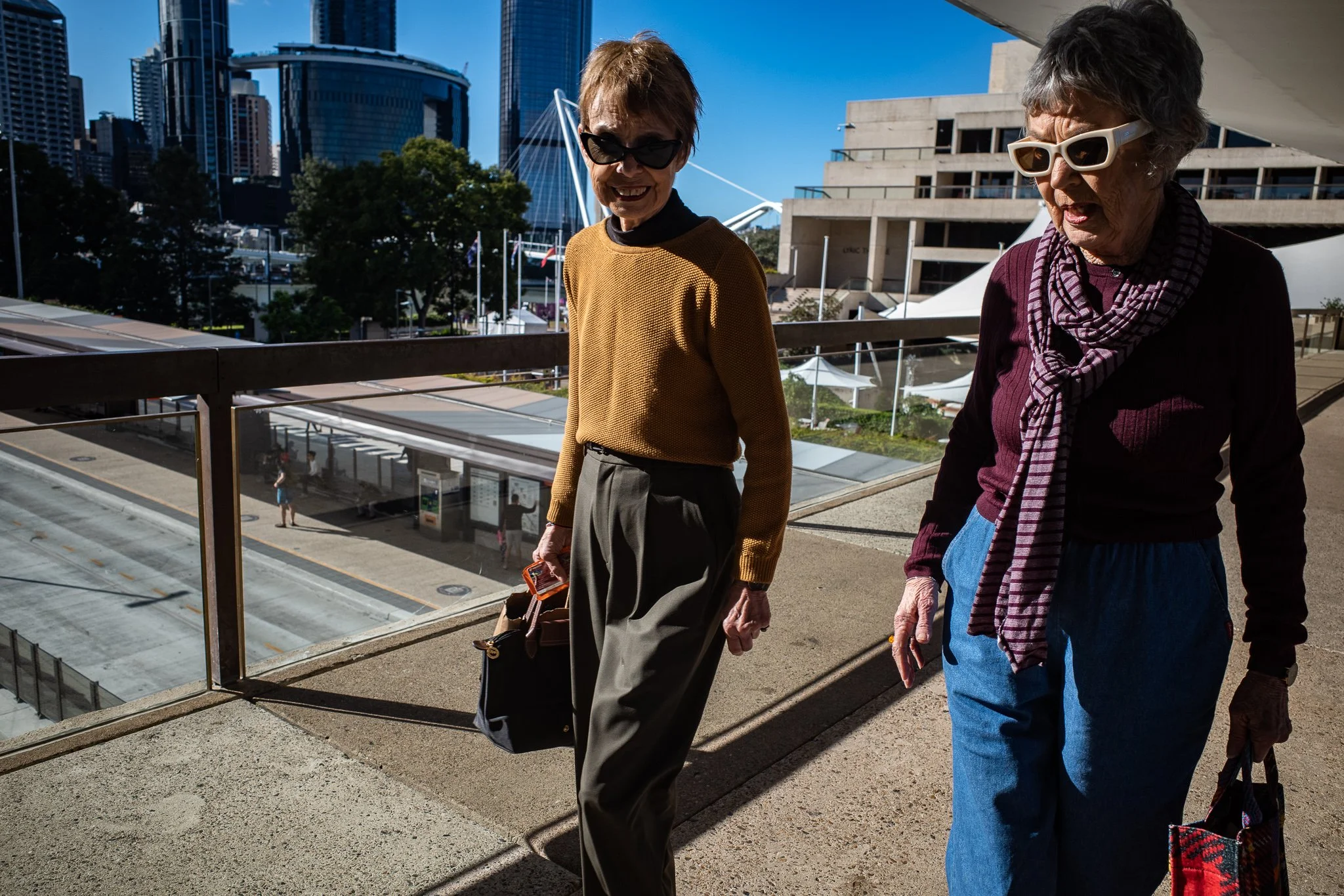 Two older women with sunglasses walking outdoors in an urban area with tall buildings in the background.