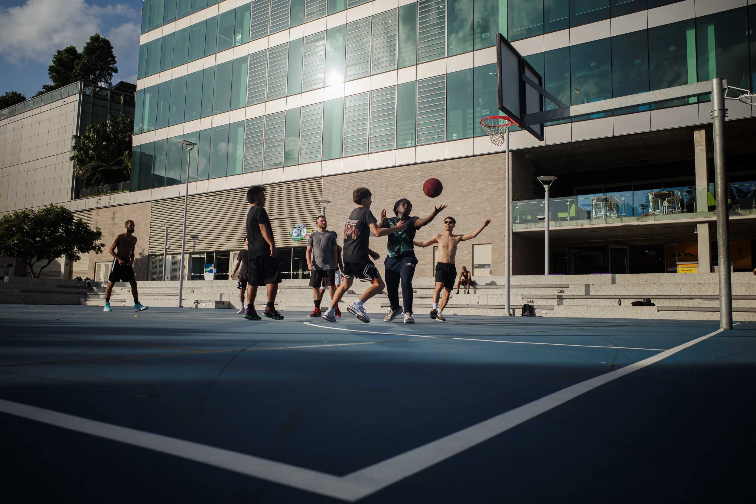 Group of young men playing basketball on an outdoor court in front of a modern glass building.