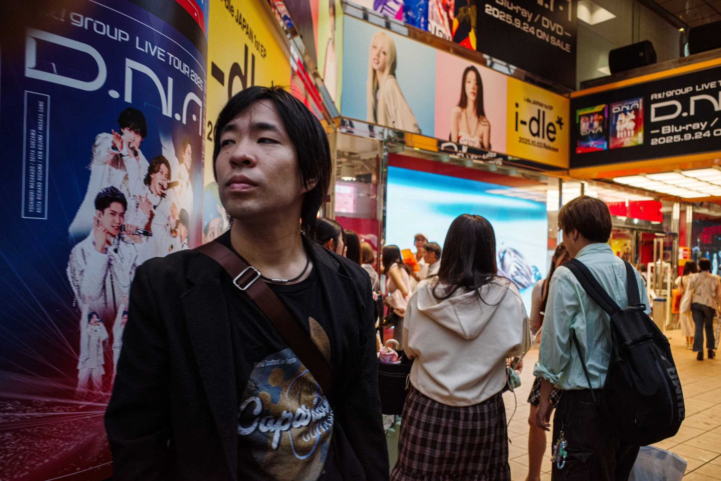 A crowded urban scene at night with people walking past brightly lit advertisements and posters, including one for a music group and another for the album i-dle.