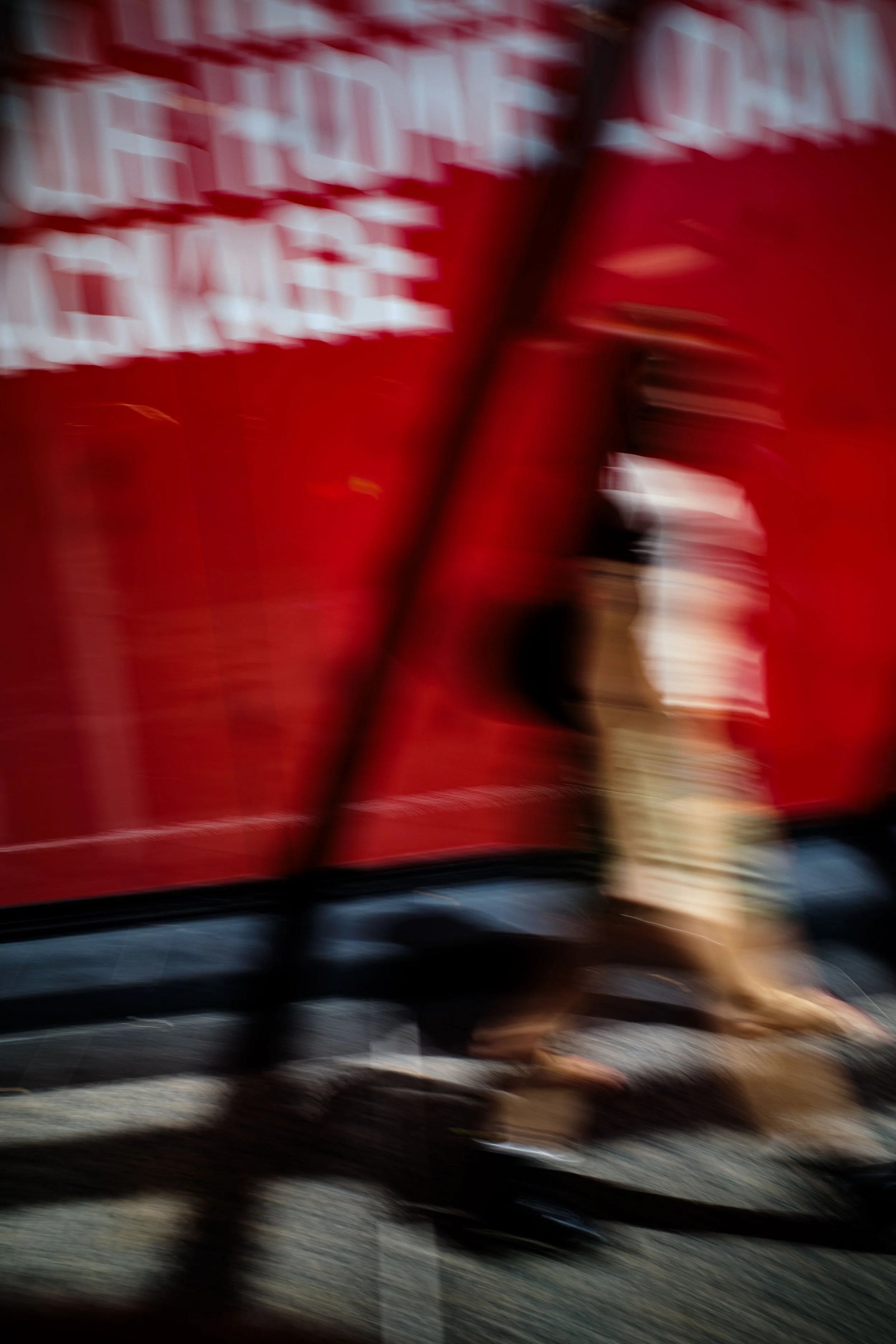 A blurred image of a person walking past a red food truck or cart with white lettering, with a black shadow or railing in the foreground.