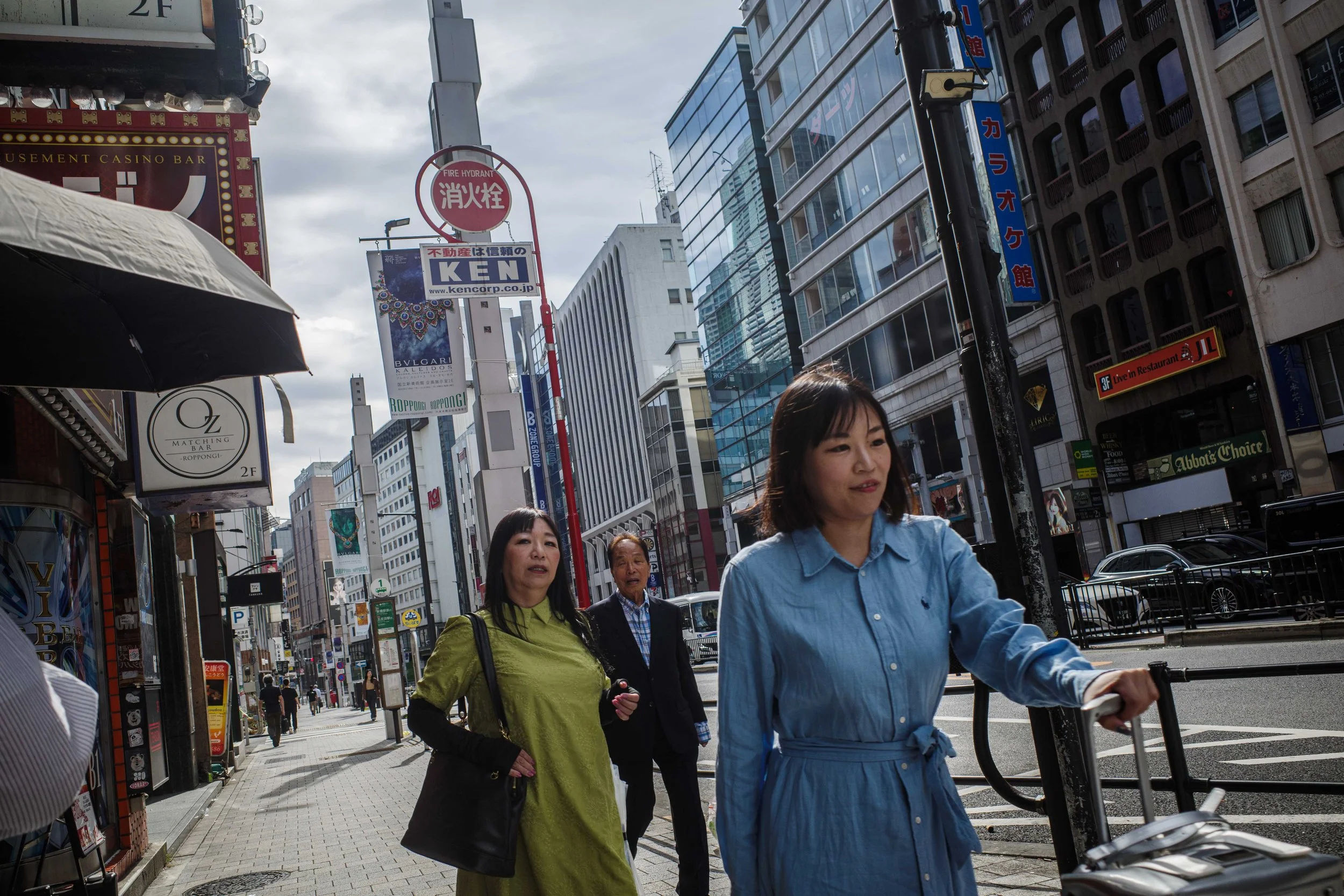 City street scene with multiple pedestrians walking along the sidewalk. Modern buildings and various signs with Japanese characters are visible. A woman in a blue dress is pushing a cart, while two other women walk behind her.