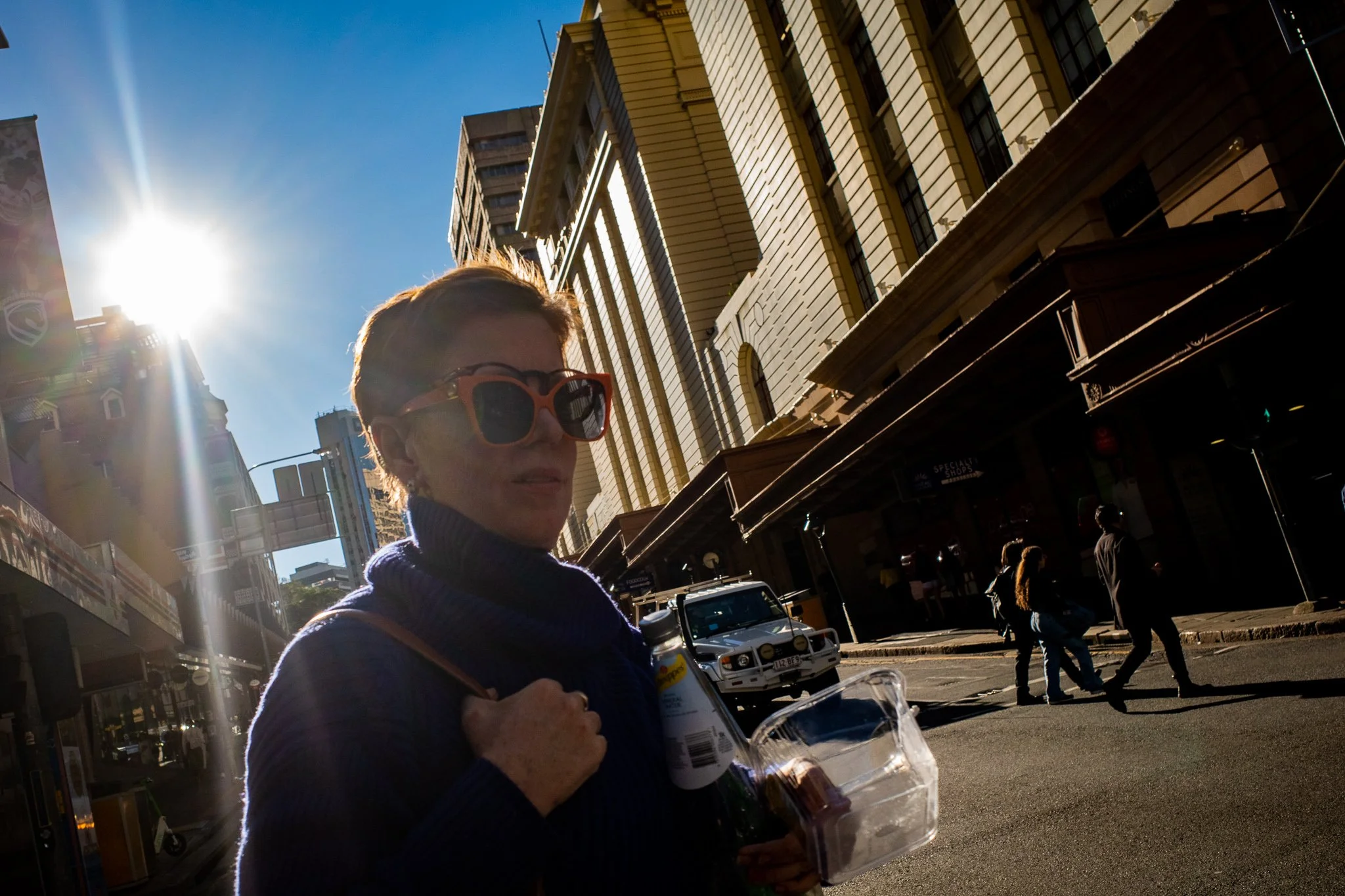 Woman wearing red sunglasses holding a drink and a plastic container walking on city street in bright sunlight, with tall buildings and pedestrians in the background.