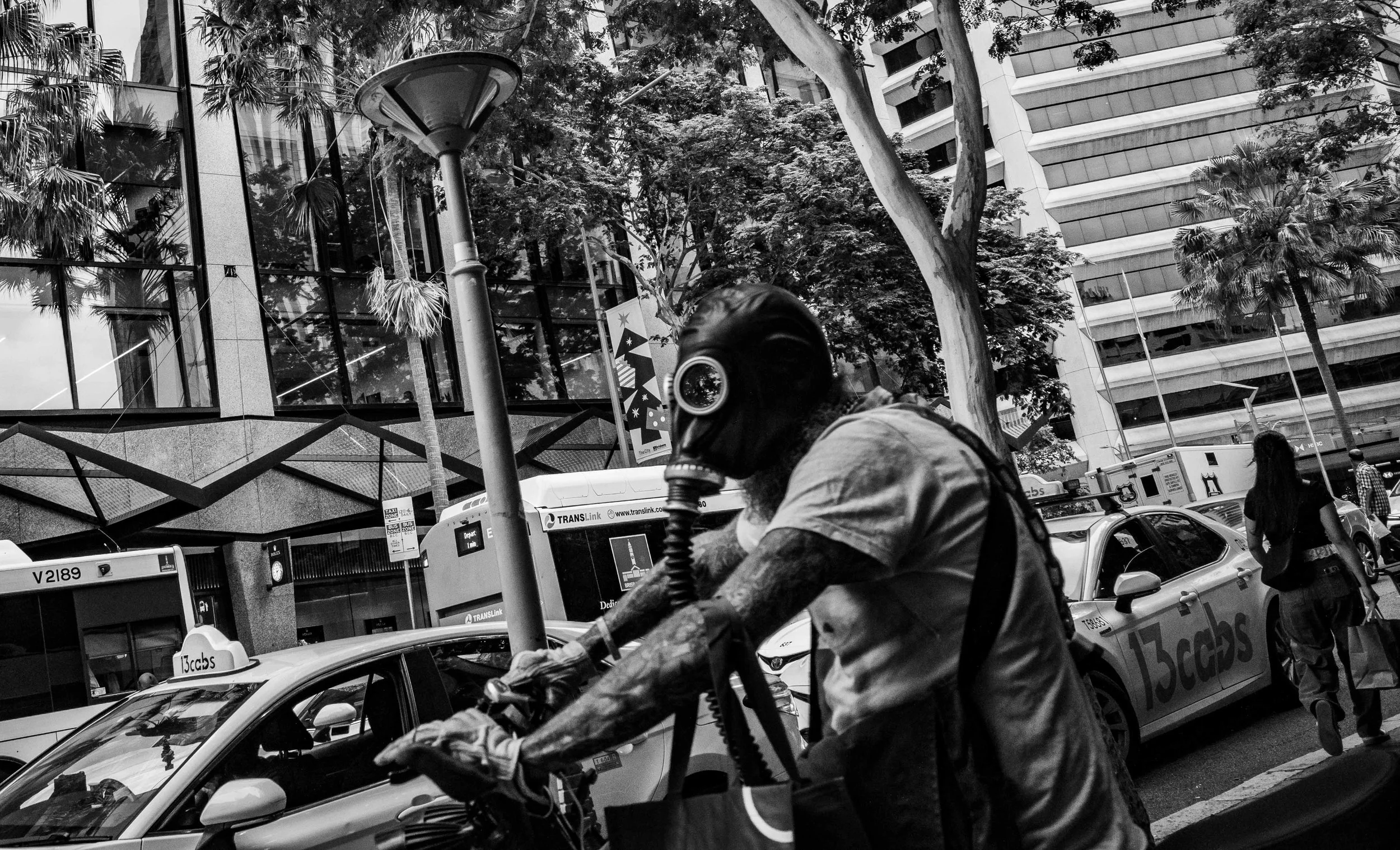 A person wearing a gas mask riding a bicycle on a busy city street, with cars and pedestrians around, high-rise buildings, and trees in the background.