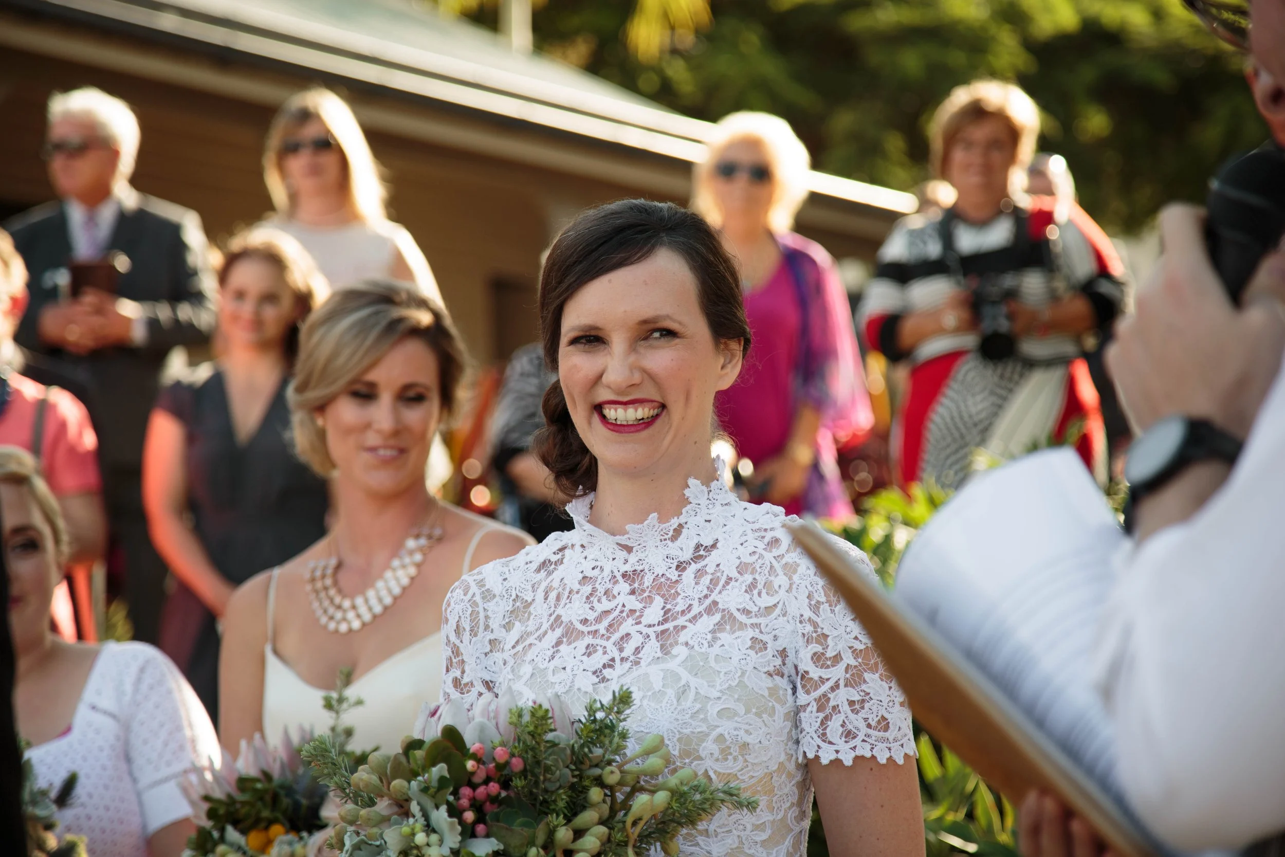 A woman in a white lace dress holding a bouquet, smiling at an outdoor wedding ceremony with guests and the officiant present.