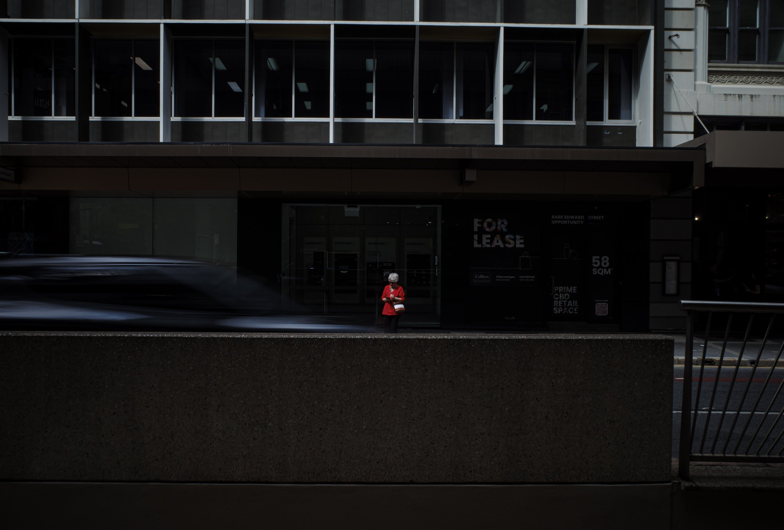 A person in a red jacket and white hair standing on a city sidewalk in front of a dark building with a 'For Lease' sign, blurred car passing by in front.