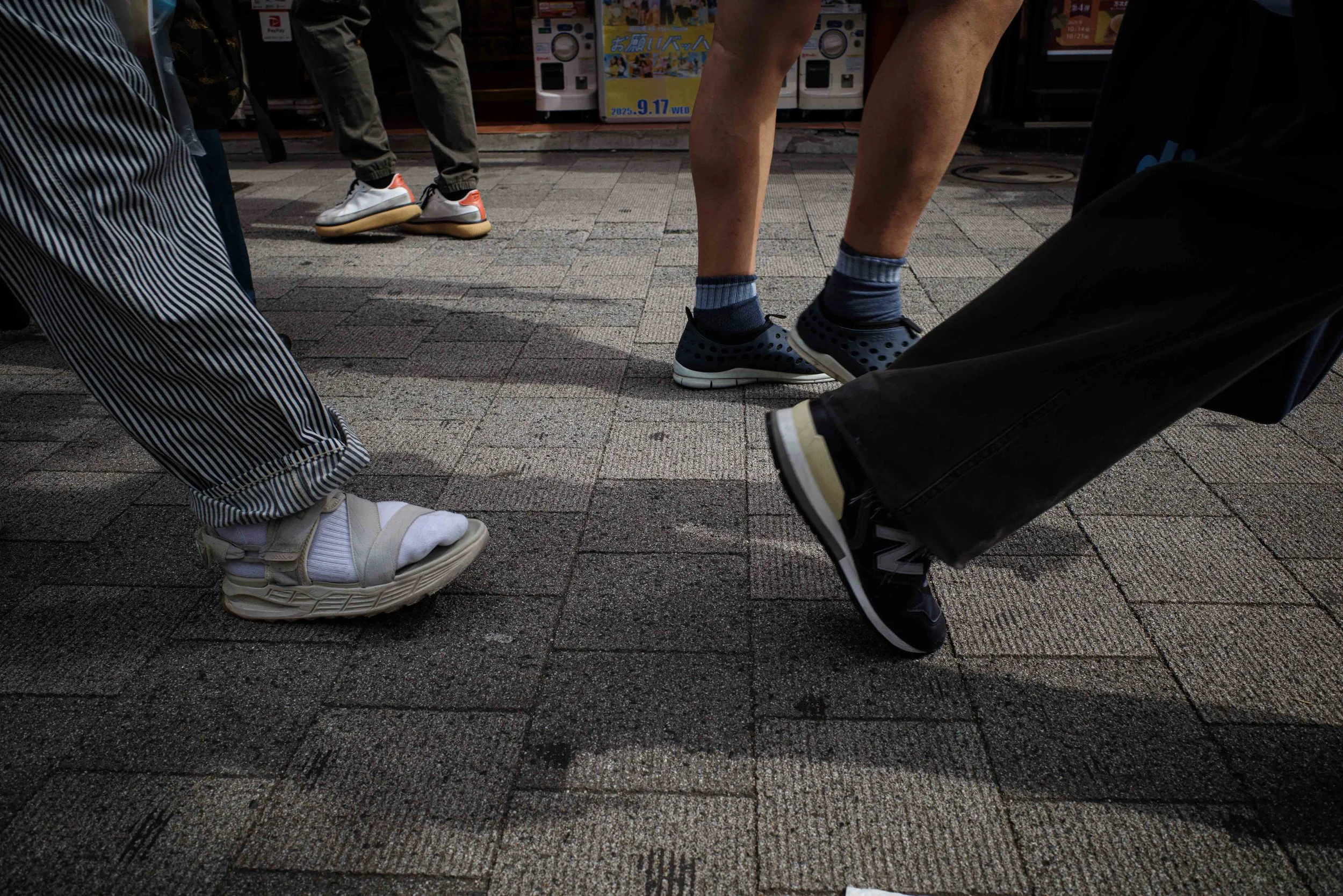 Close-up of several people’s feet and legs, with some dressed in sneakers and others in sandals, crossing or moving on a paved sidewalk in an urban area.