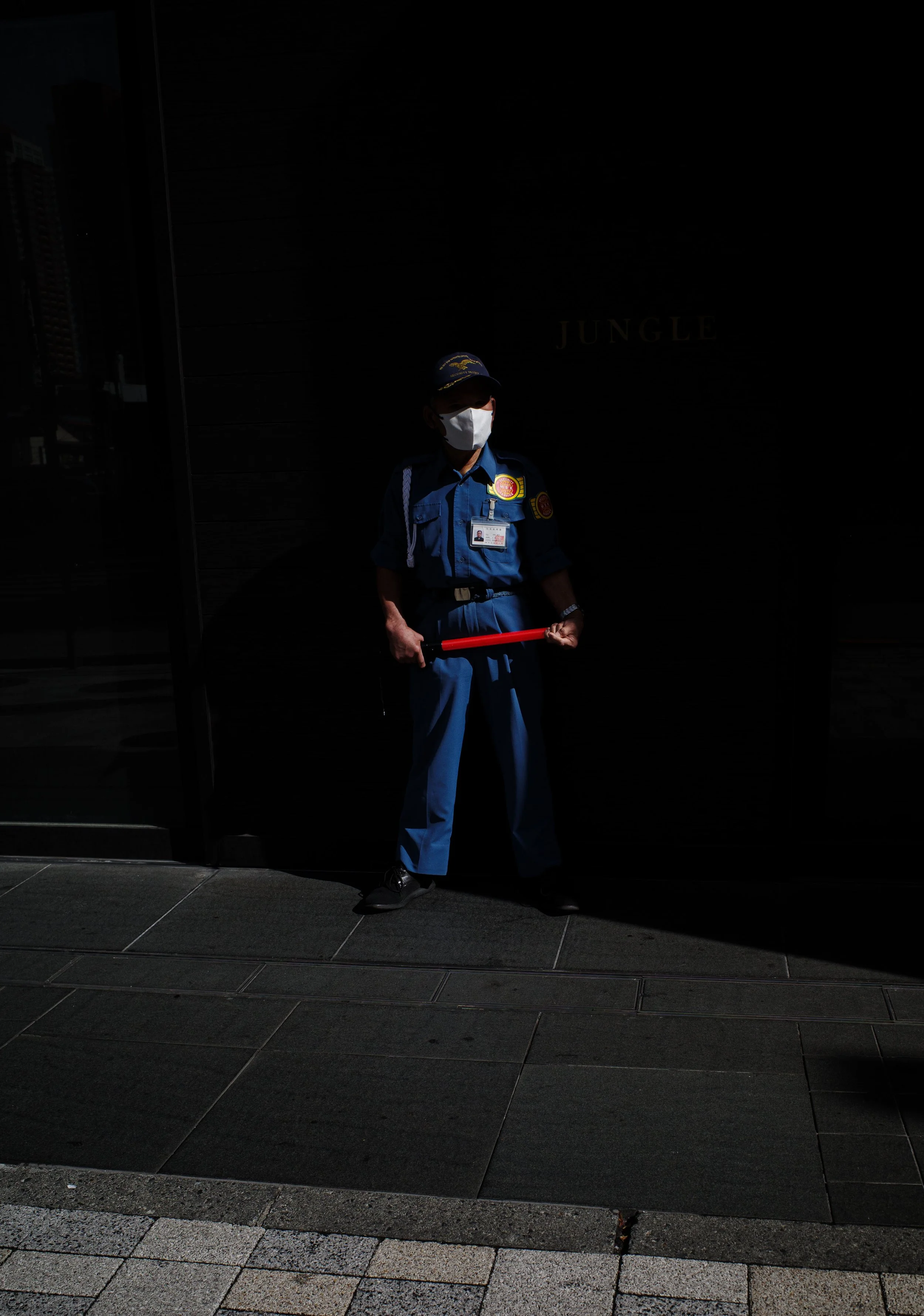 A security guard wearing a face mask and a blue uniform stands in a shadowed area, holding a red baton.