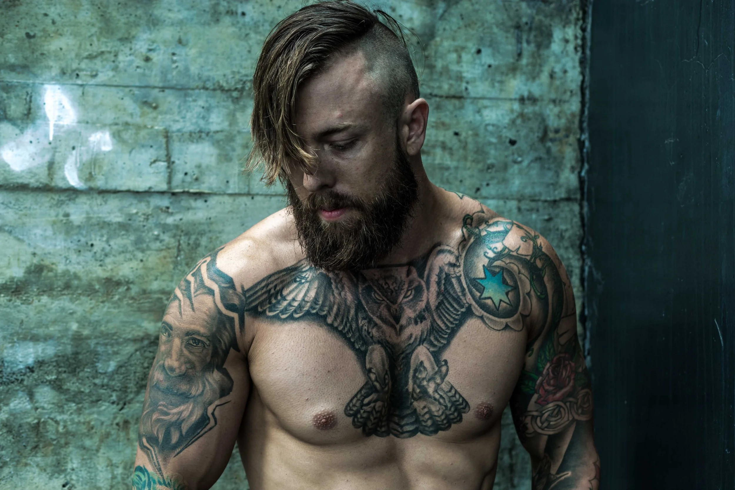 A shirtless man with tattoos on his chest and arms, with a beard and long, partially shaved hair, standing against a weathered green wall.