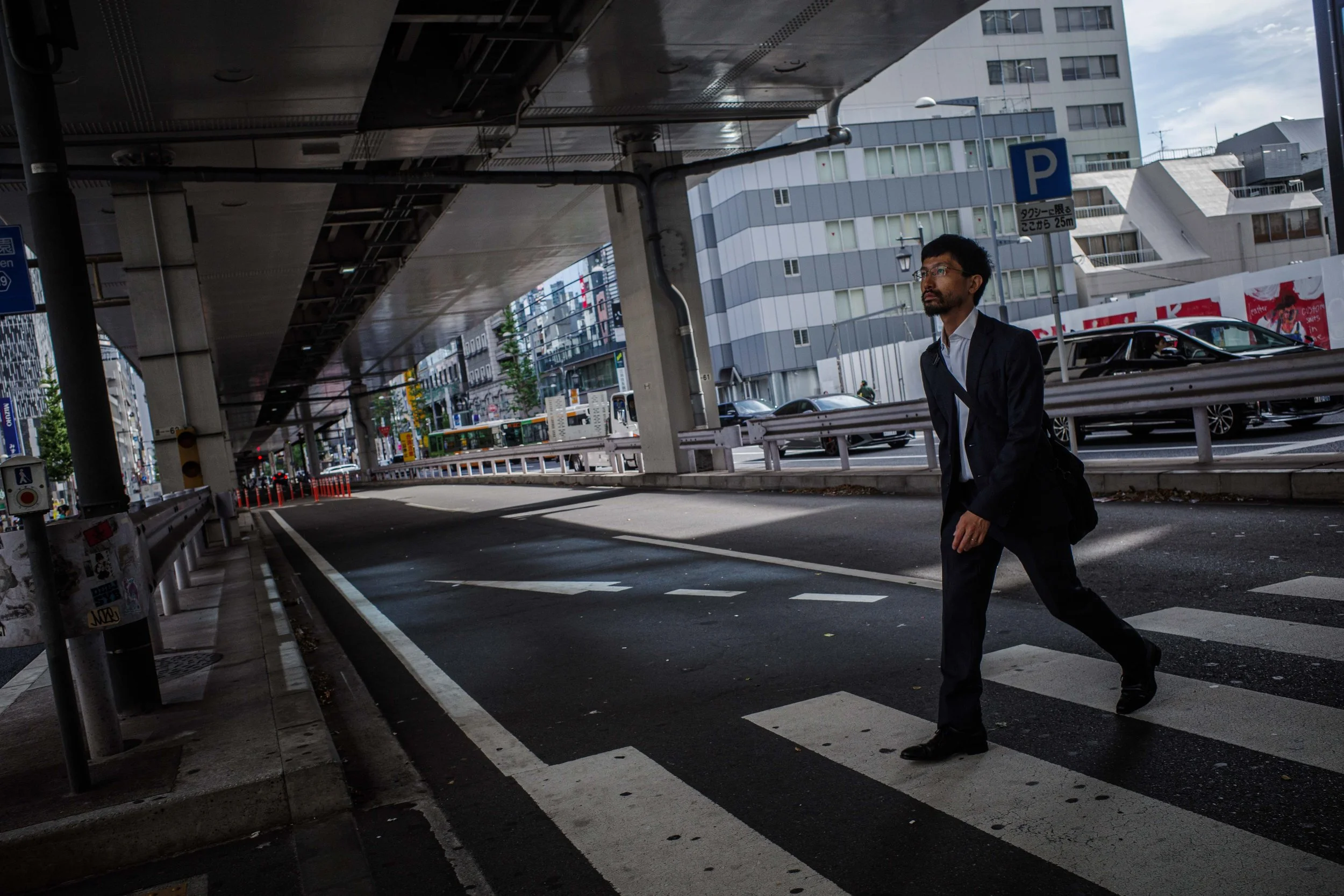 A man in a dark suit walks across a crosswalk under an overpass in an urban area.