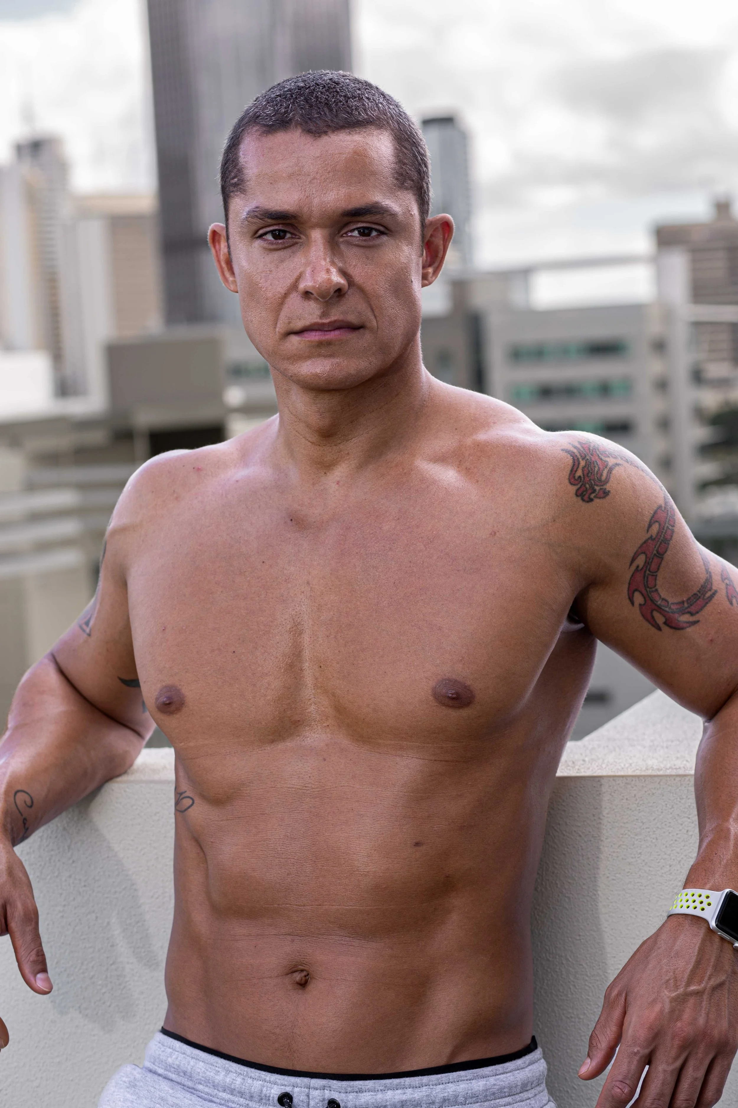 A shirtless man with short dark hair and tattoos on his arms and shoulder, standing outdoors on a city rooftop with a skyline of tall buildings in the background.