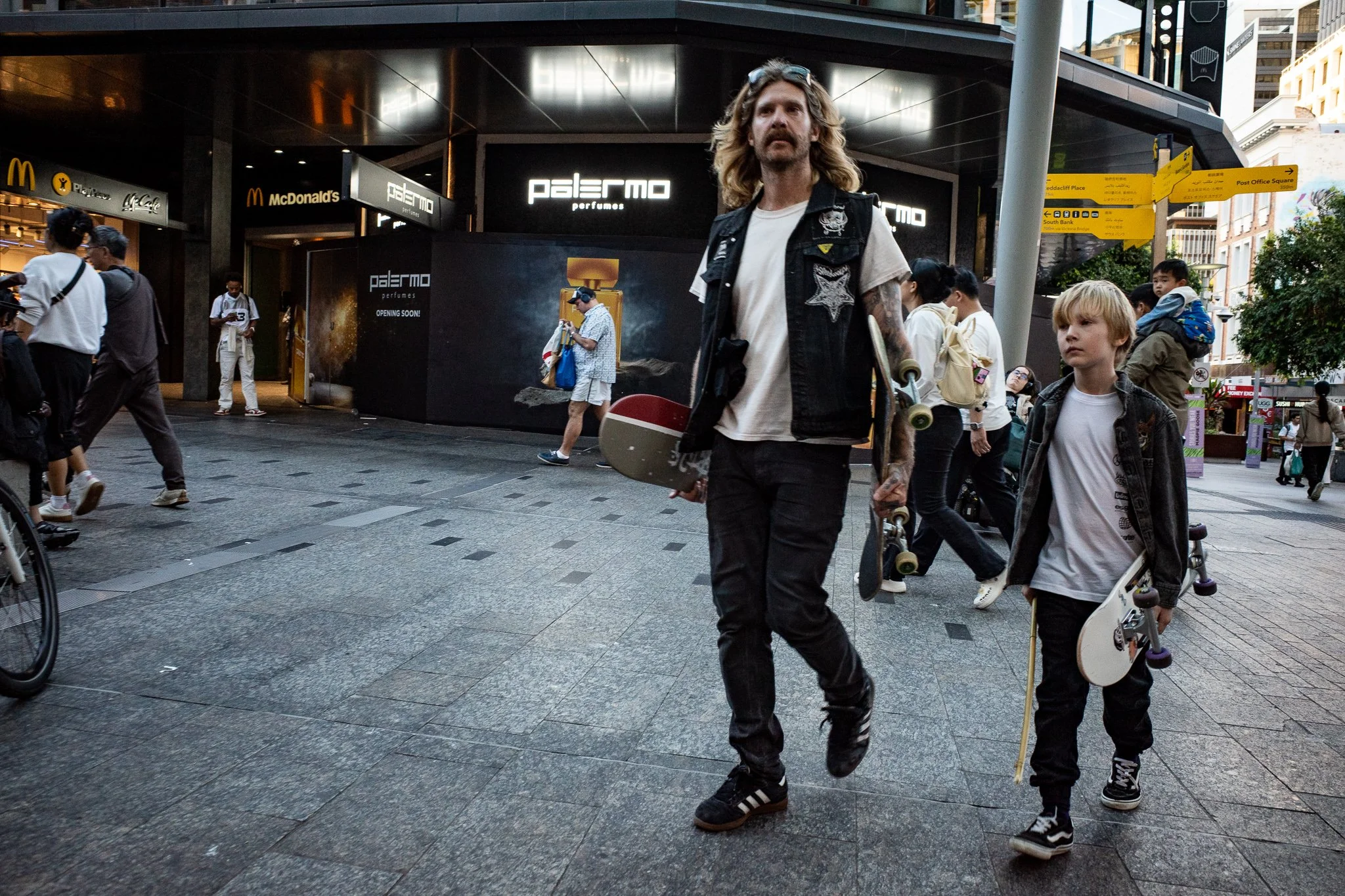 Street scene with pedestrians, including a man with long hair and a skateboard and a boy with blonde hair and a skateboard, in front of shops and signs, including a perfume store named Palermo and a McDonald's.