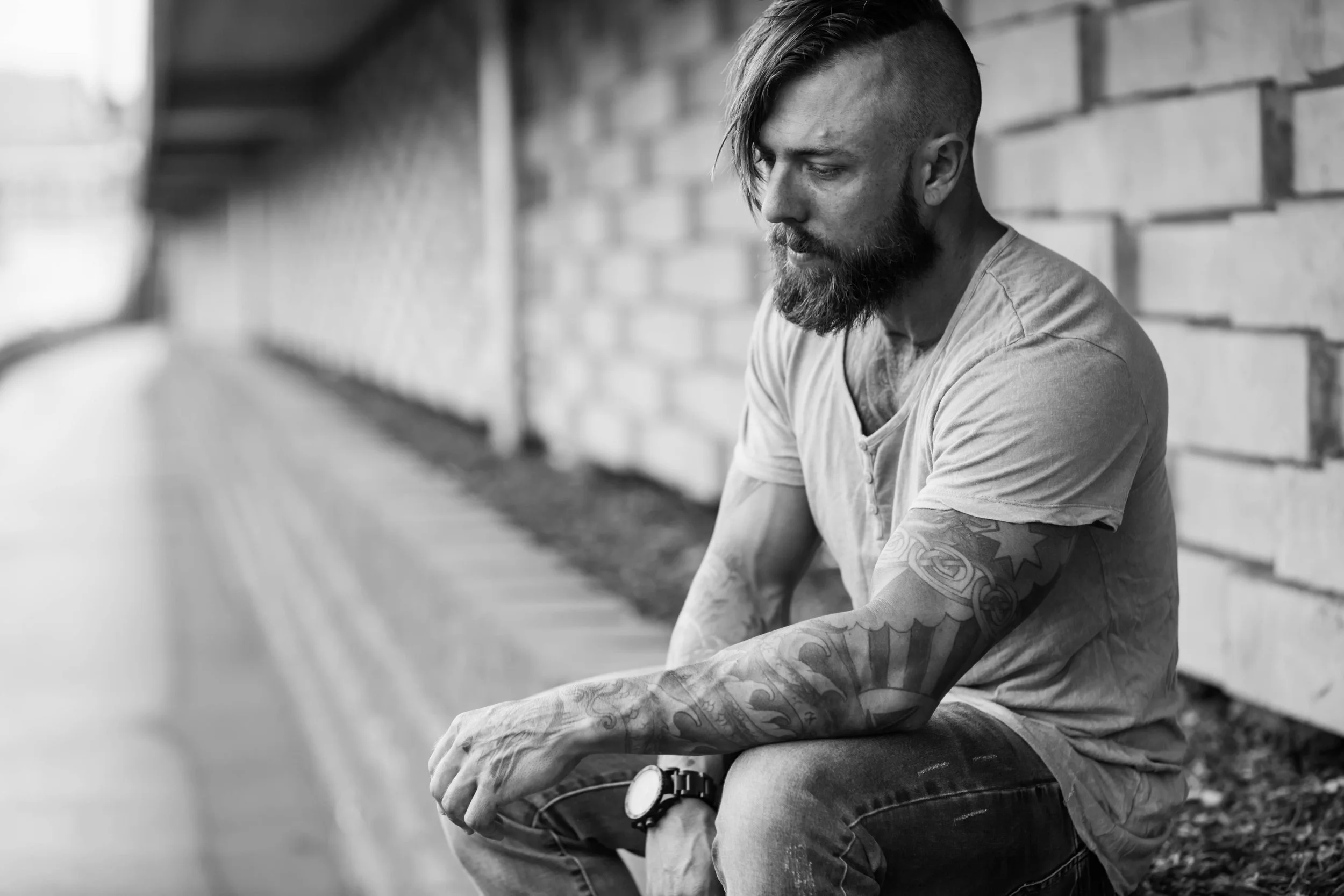 A man with tattoos and a beard sitting by a brick wall, looking down thoughtfully.