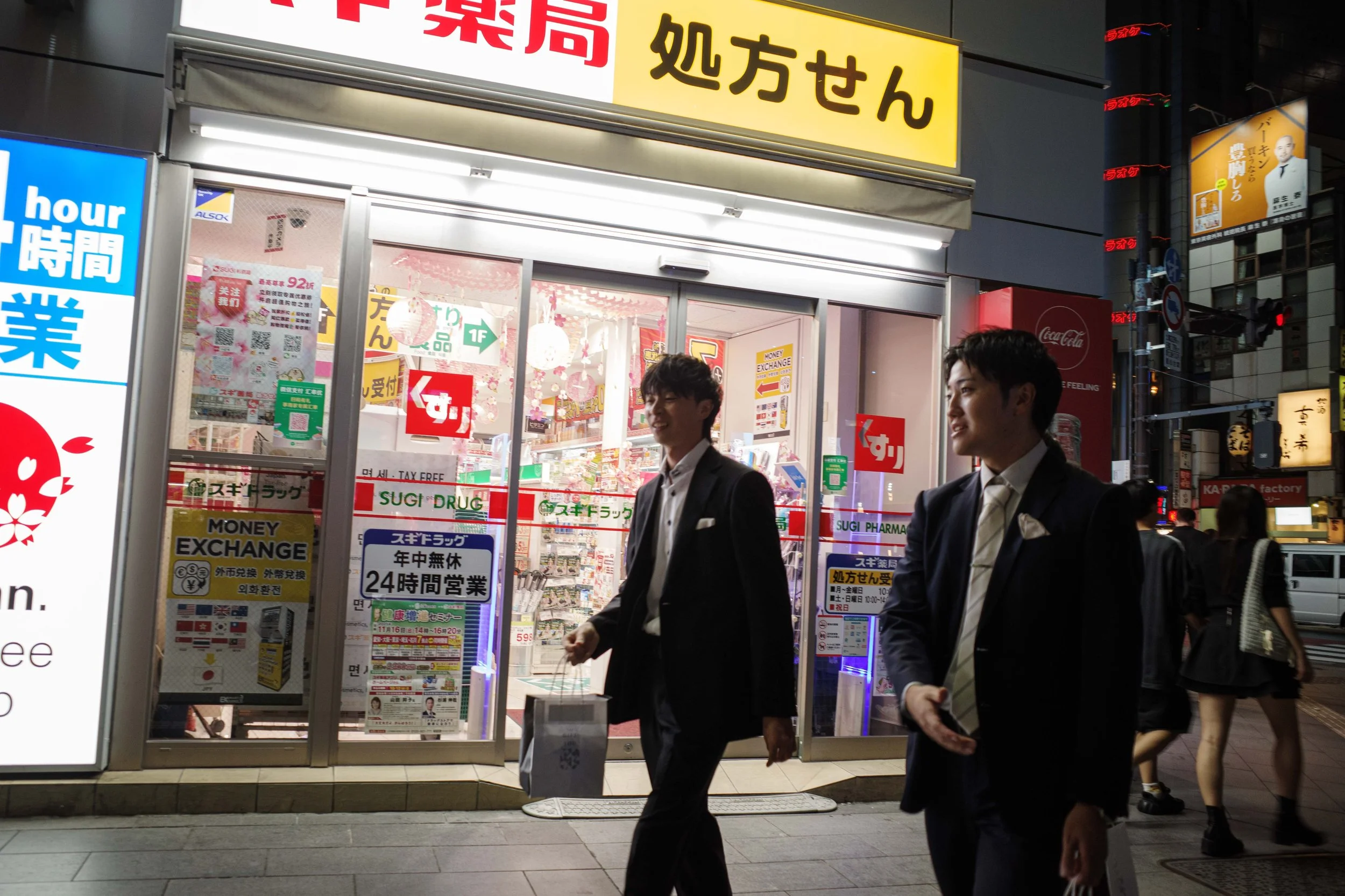 Nighttime street scene with two young men in suits walking past a pharmacy store with bright signage, glass doors, and colorful displays.