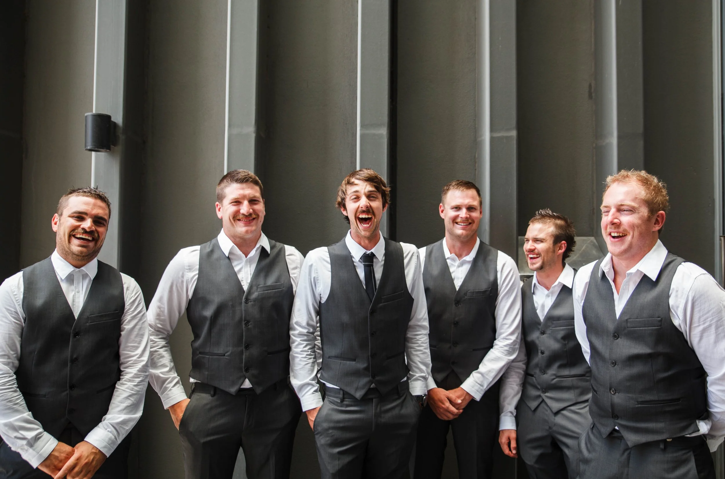Group of six young men in gray vests and white shirts, smiling and laughing in front of a modern gray wall.