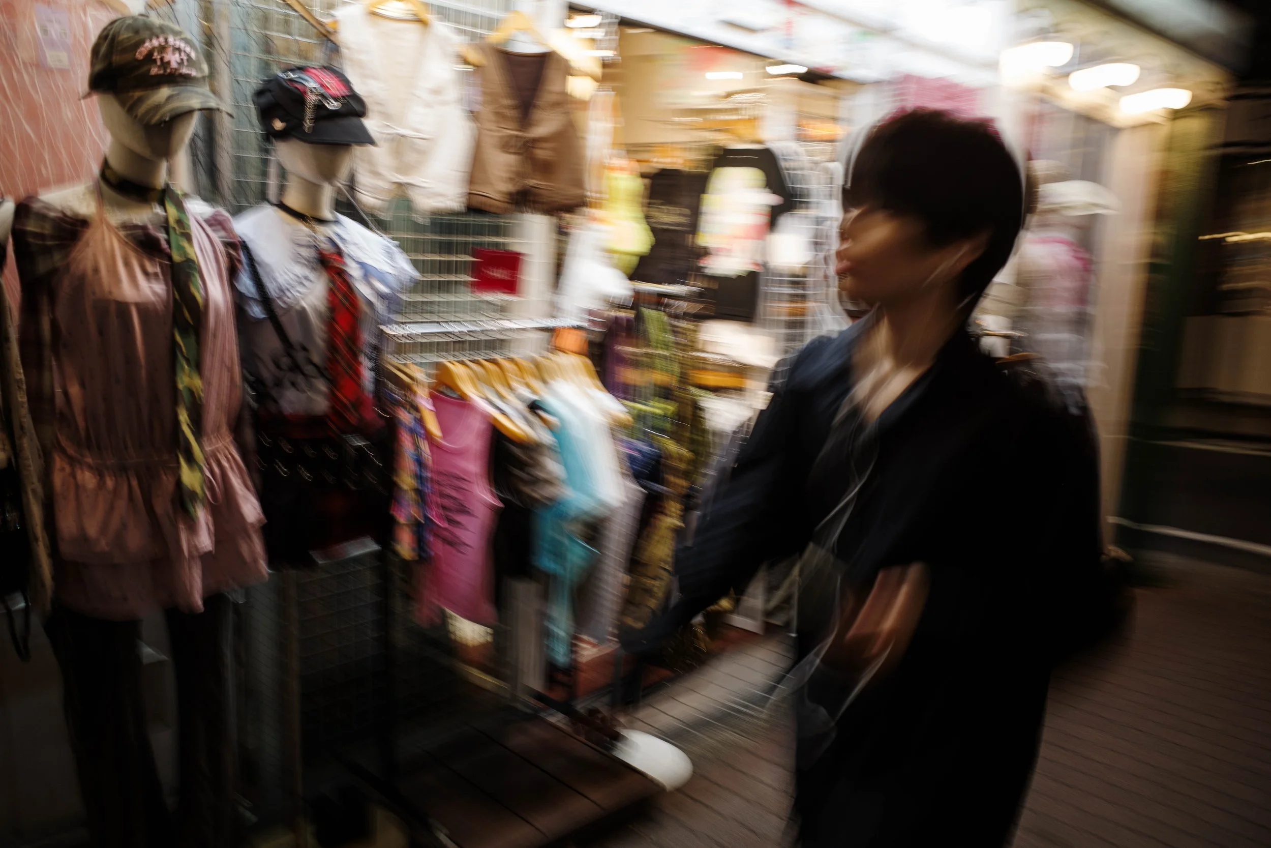 A woman shopping on a street, looking at mannequins dressed in clothing, with colorful outfits and hats, in an outdoor market stall.