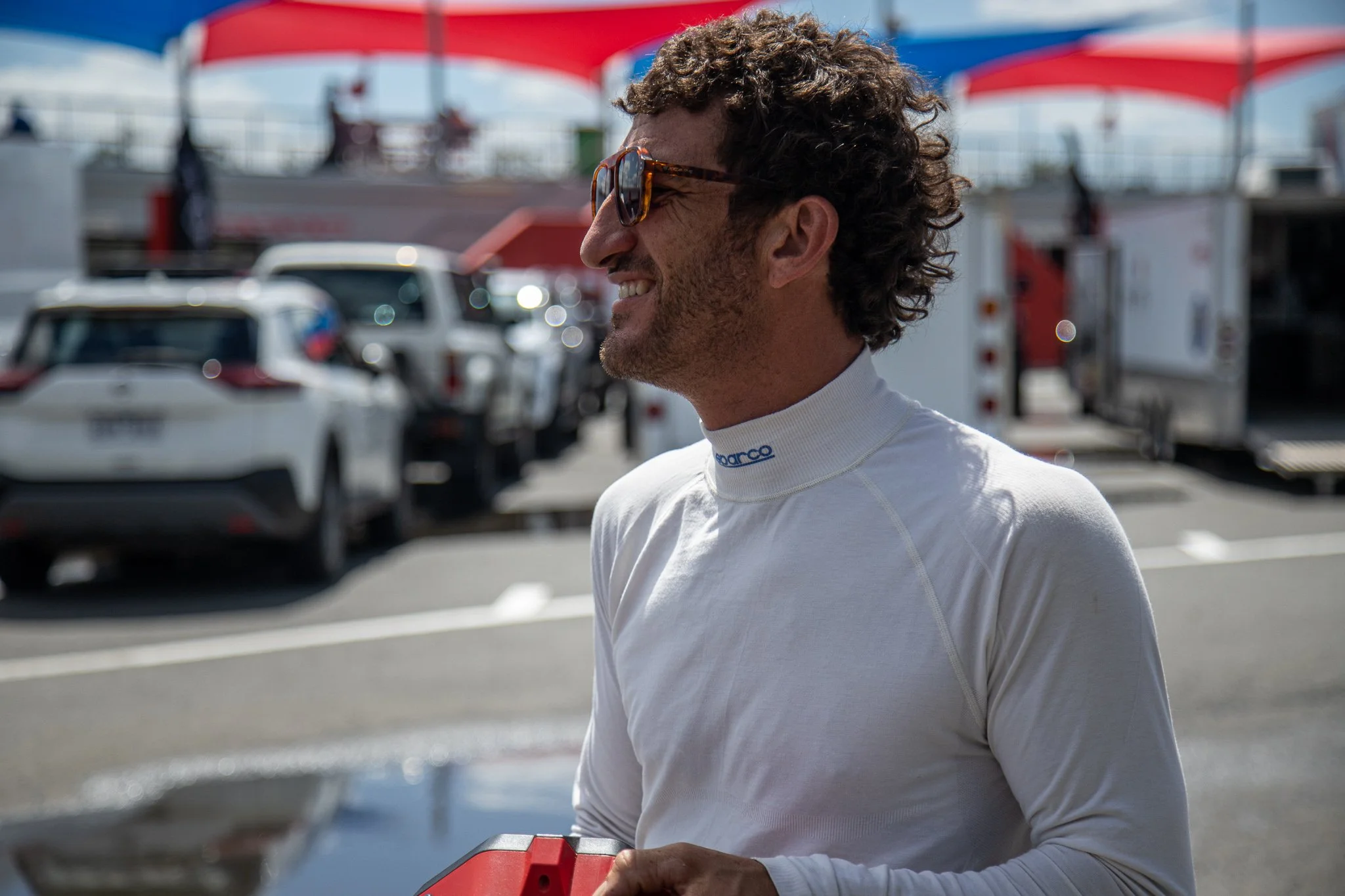 A man with curly hair, wearing sunglasses and a white racing suit, smiling in a racing paddock area with cars and equipment in the background on a sunny day.