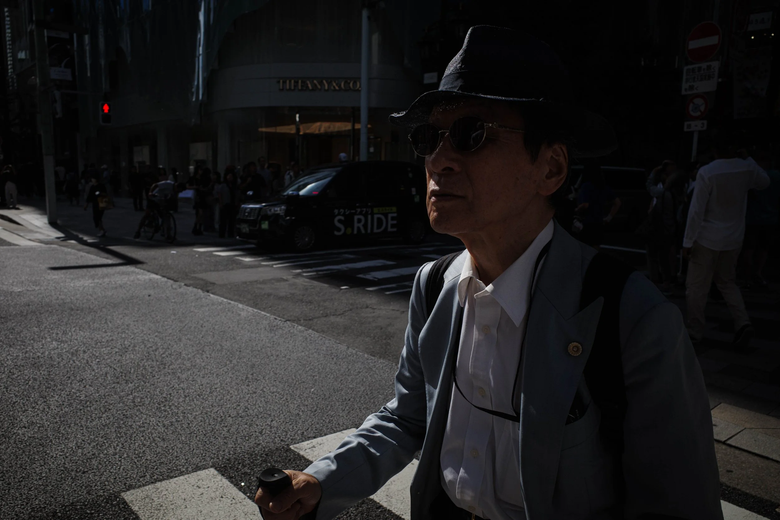 A man wearing sunglasses, a hat, and a suit walking across a city street on a sunny day.