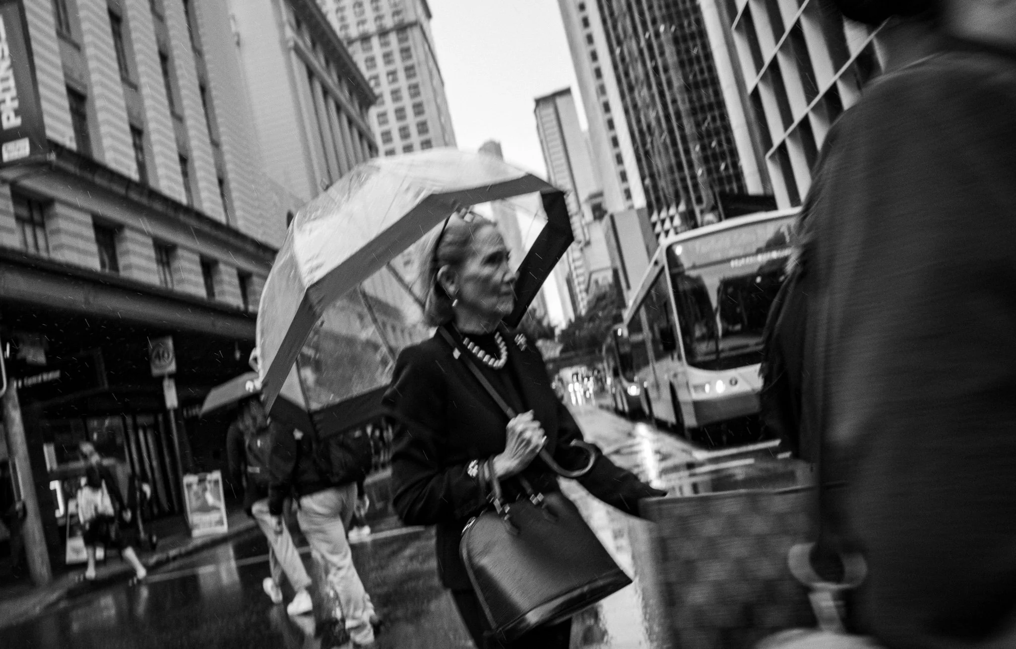 A woman holding an umbrella walking on a busy city street next to a person with a case, with tall buildings and a bus in the background.