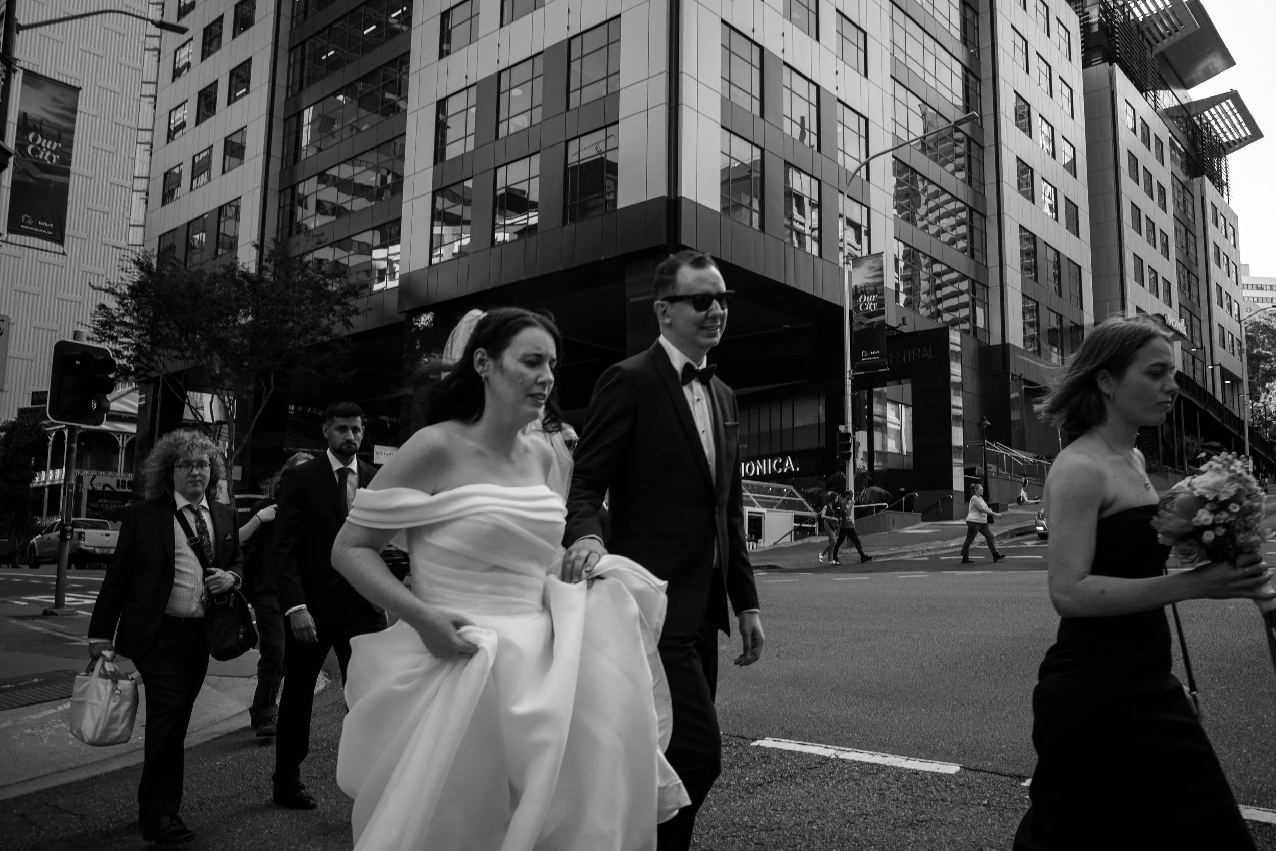 Black and white photo of a wedding scene on a city street with a bride in a wedding dress, and a groom in a tuxedo with sunglasses, walking together with bridesmaids and guests.