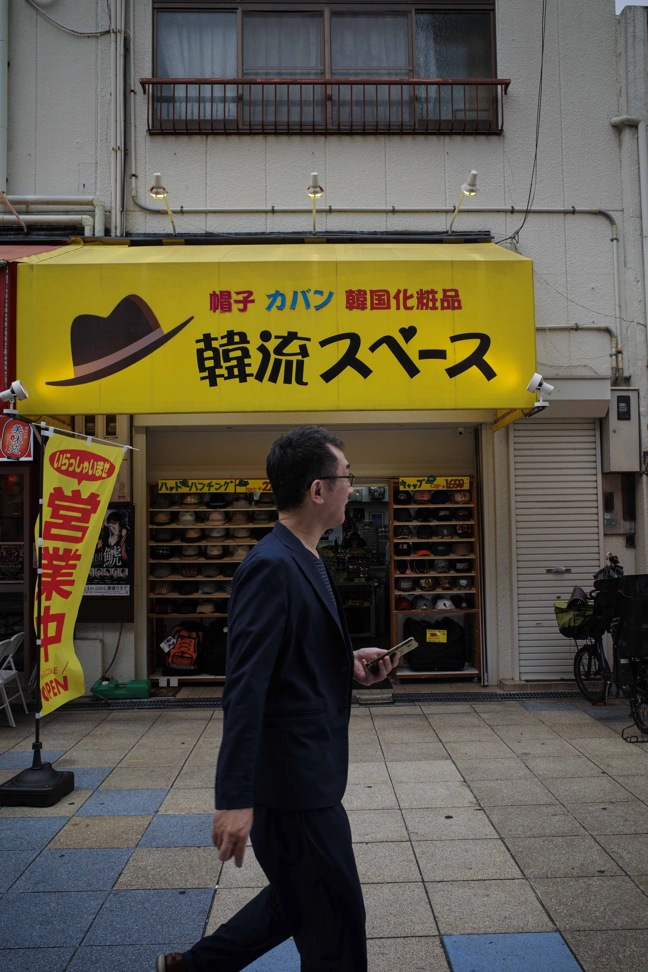 A man in a navy suit walking past a store with a yellow sign in Japanese, selling souvenirs and Korean cosmetics, with hats displayed outside.