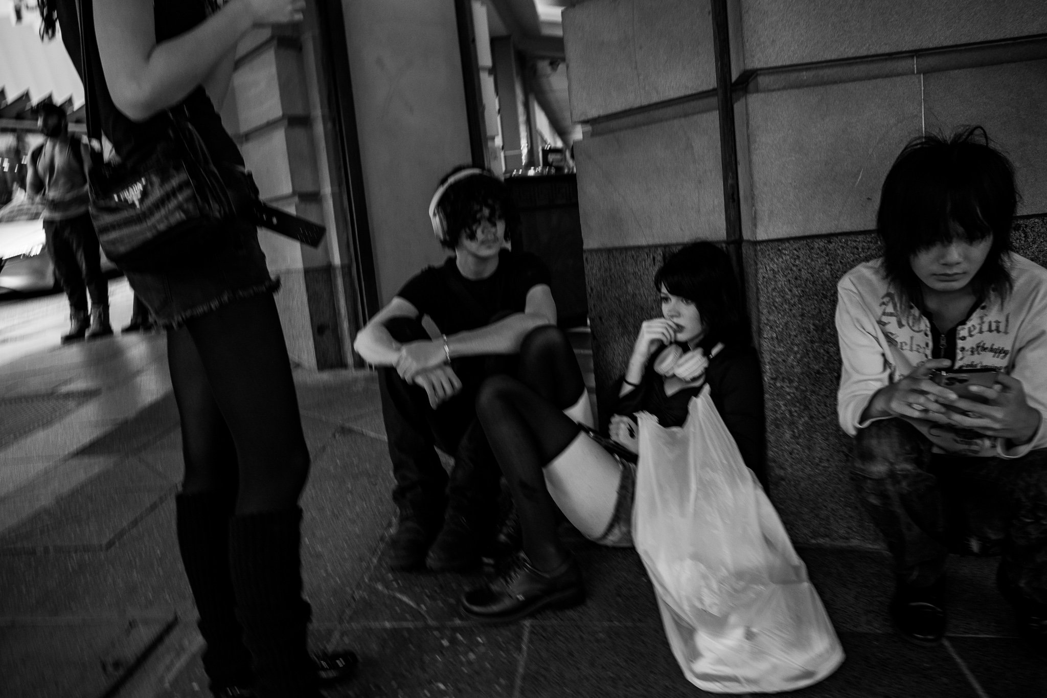 Three young people sitting on the sidewalk against a building wall, with a woman standing nearby. Two of them are looking at their phones, and one has headphones around her neck. A shopping bag is on the ground beside them.