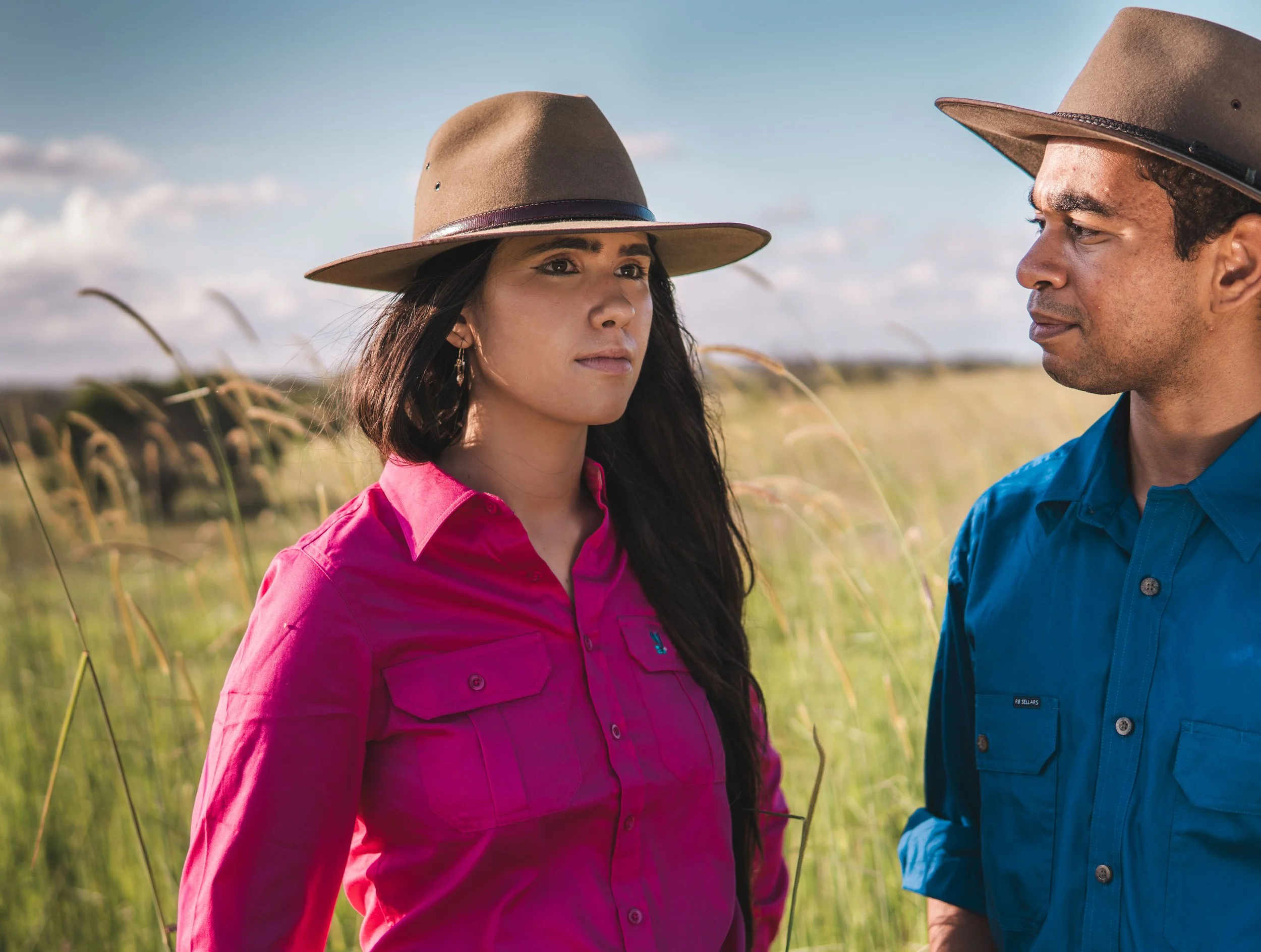 A woman and a man wearing hats and outdoor clothing standing in a field with tall grass and a partly cloudy sky.