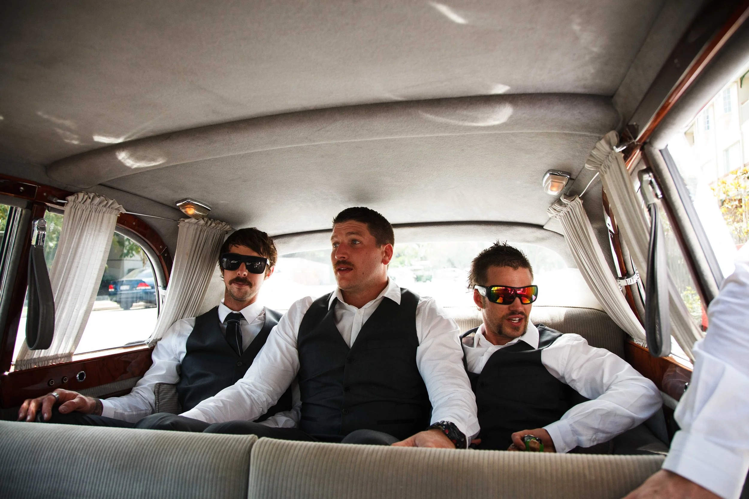 Three men in formal attire and sunglasses sitting inside a vintage car, possibly a limousine, with white curtains on the windows.