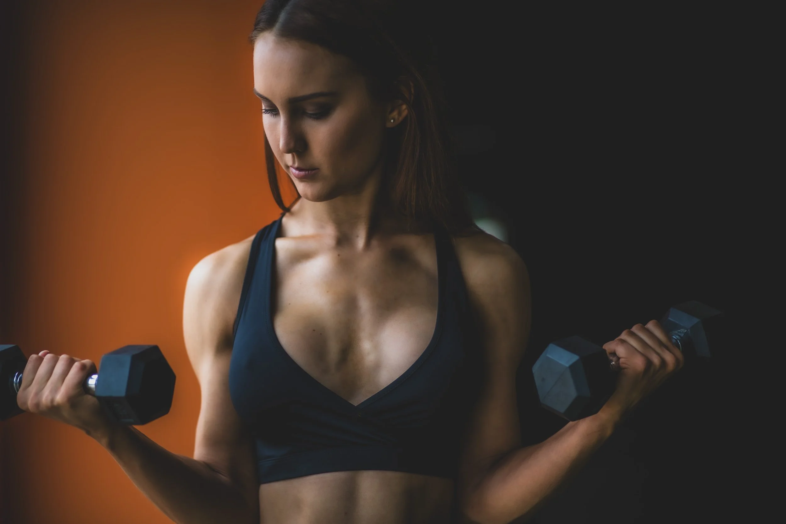 A woman in athletic wear lifts dumbbells in a gym, with a dark background and a warm orange light on her left side.