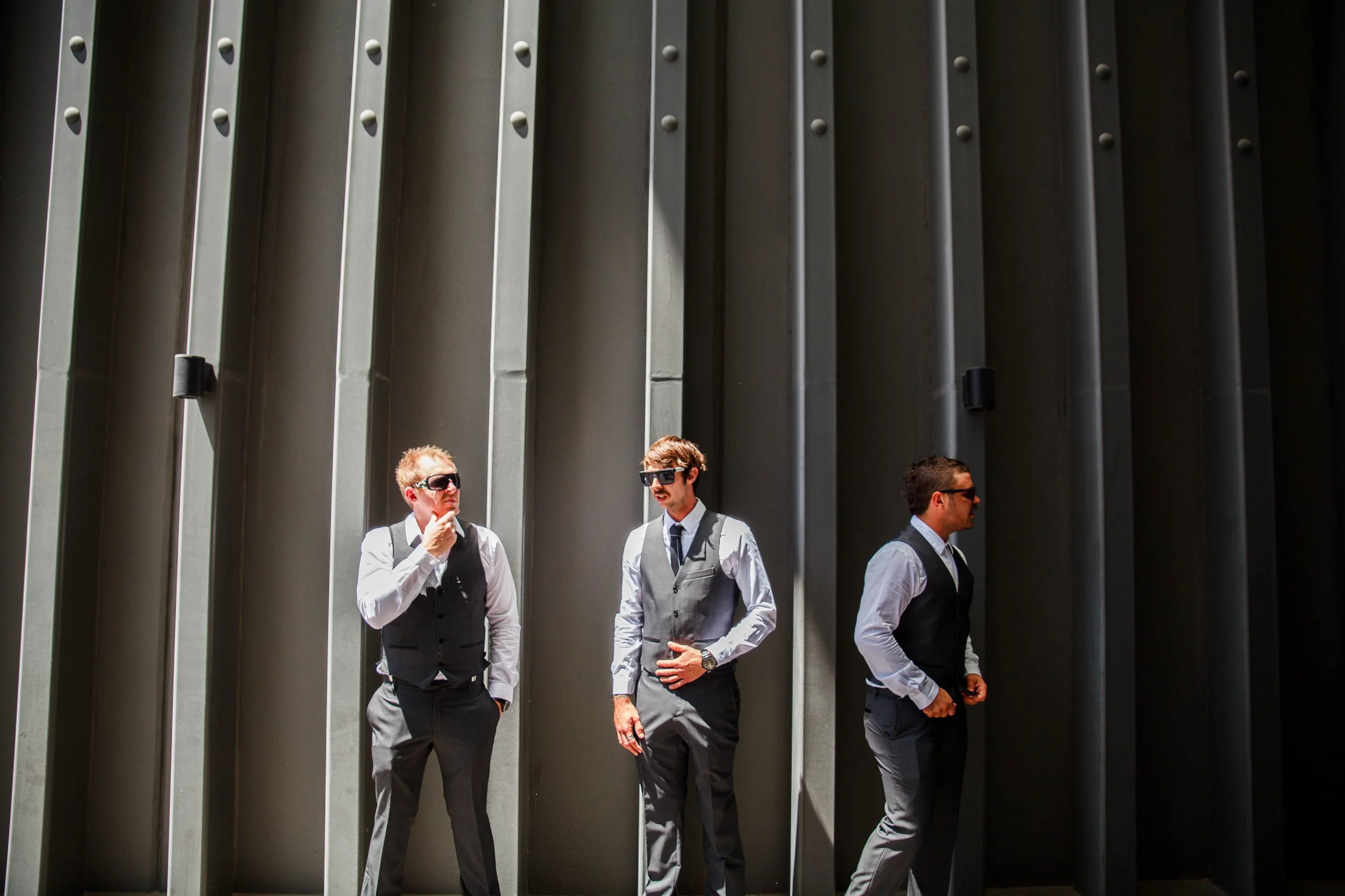 Three men in business attire standing in front of a tall, dark, metal wall, wearing sunglasses and engaged in thought or conversation.