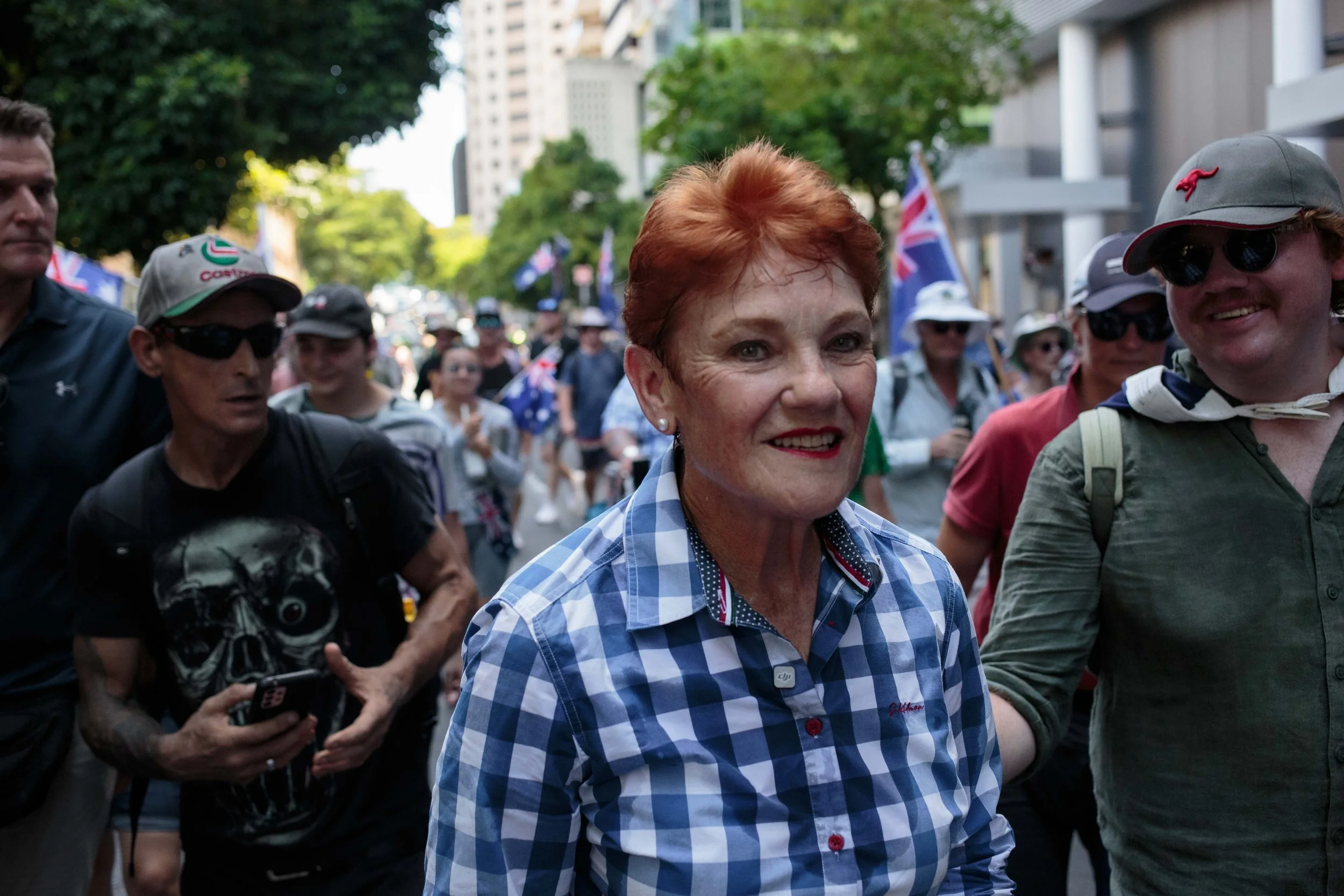 A group of people participating in a parade or march, with a woman with short red hair and wearing a blue and white checkered shirt smiling at the front. Others around her are wearing hats and sunglasses, and Australian flags are visible in the backg