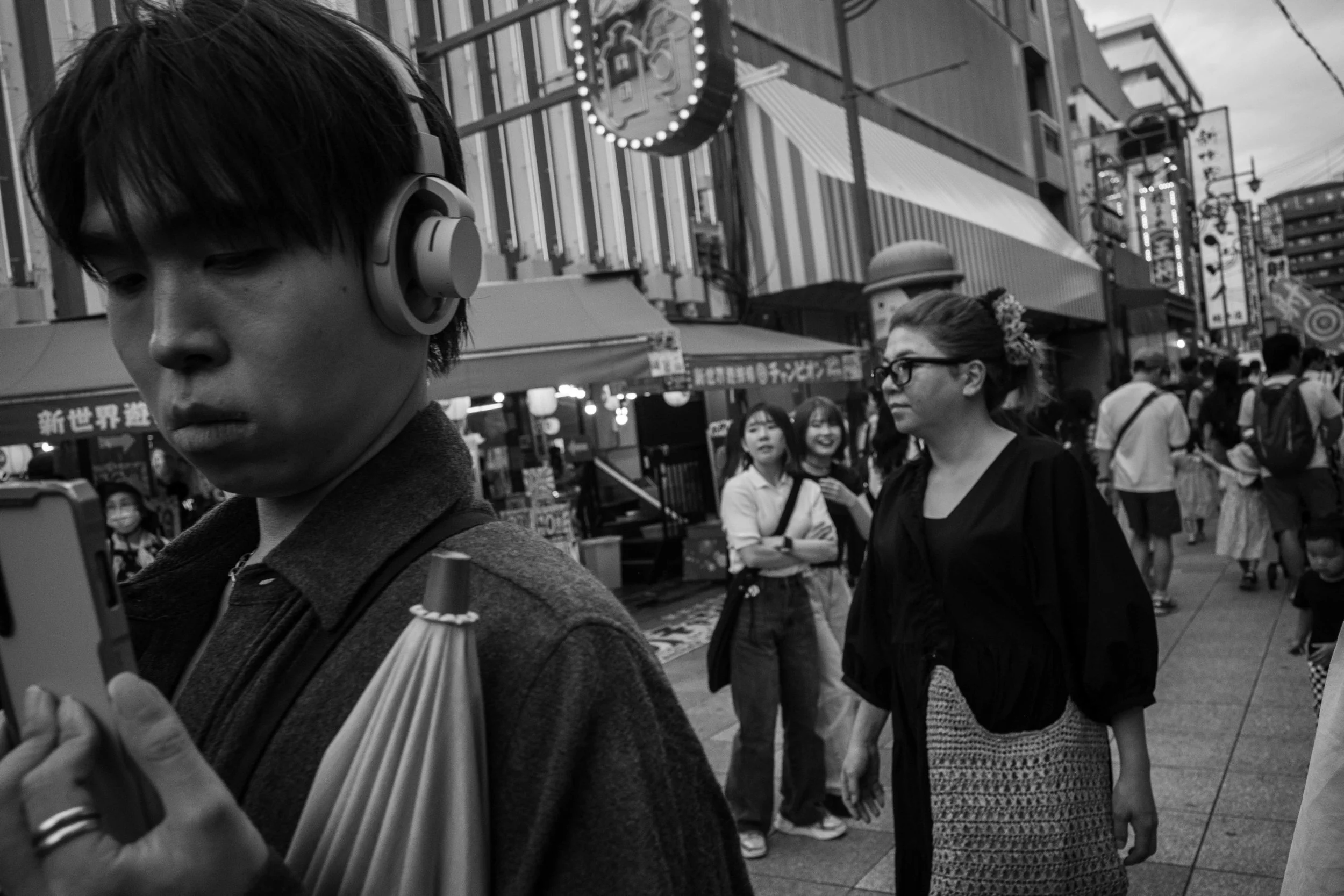 A black and white photo of a busy street scene with several people. The foreground features a young man with headphones looking at his phone, with a person in the background looking to the side and laughing.