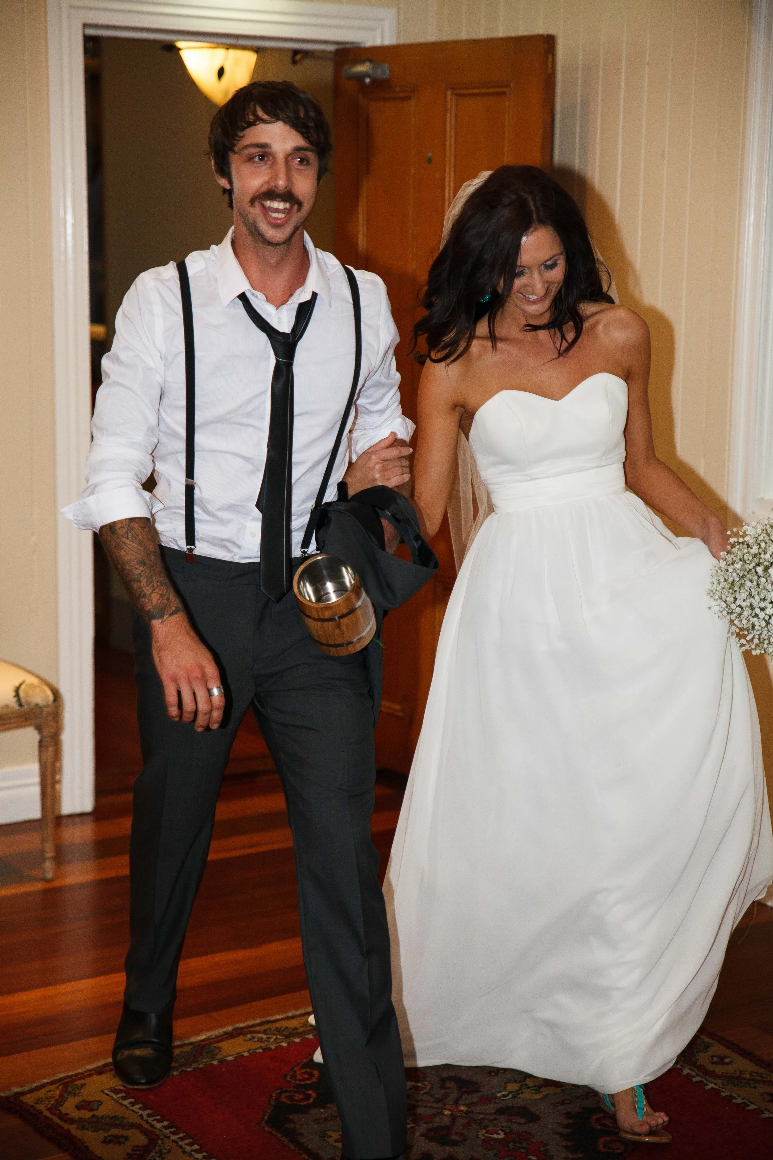 A man and a woman, possibly bride and groom, walking together indoors. The woman is wearing a white strapless wedding gown and holding a bouquet, while the man is dressed in formal attire with suspenders, a tie, and tattoos visible on his arm.