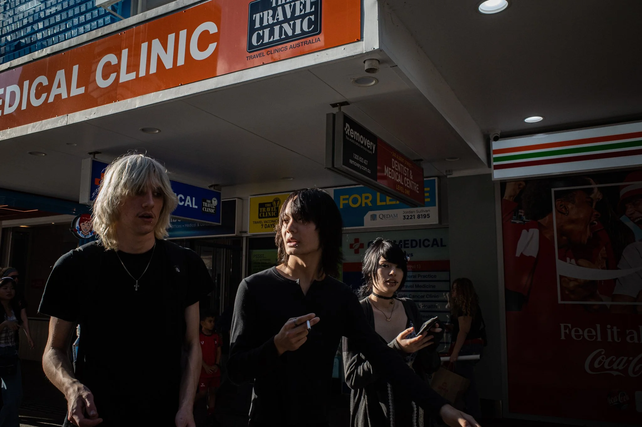 People walking in front of a medical clinic with signs and posters, including a large orange sign that reads 'MEDICAL CLINIC' and smaller signs advertising dental and medical services, with a lineup of individuals in dark clothing.