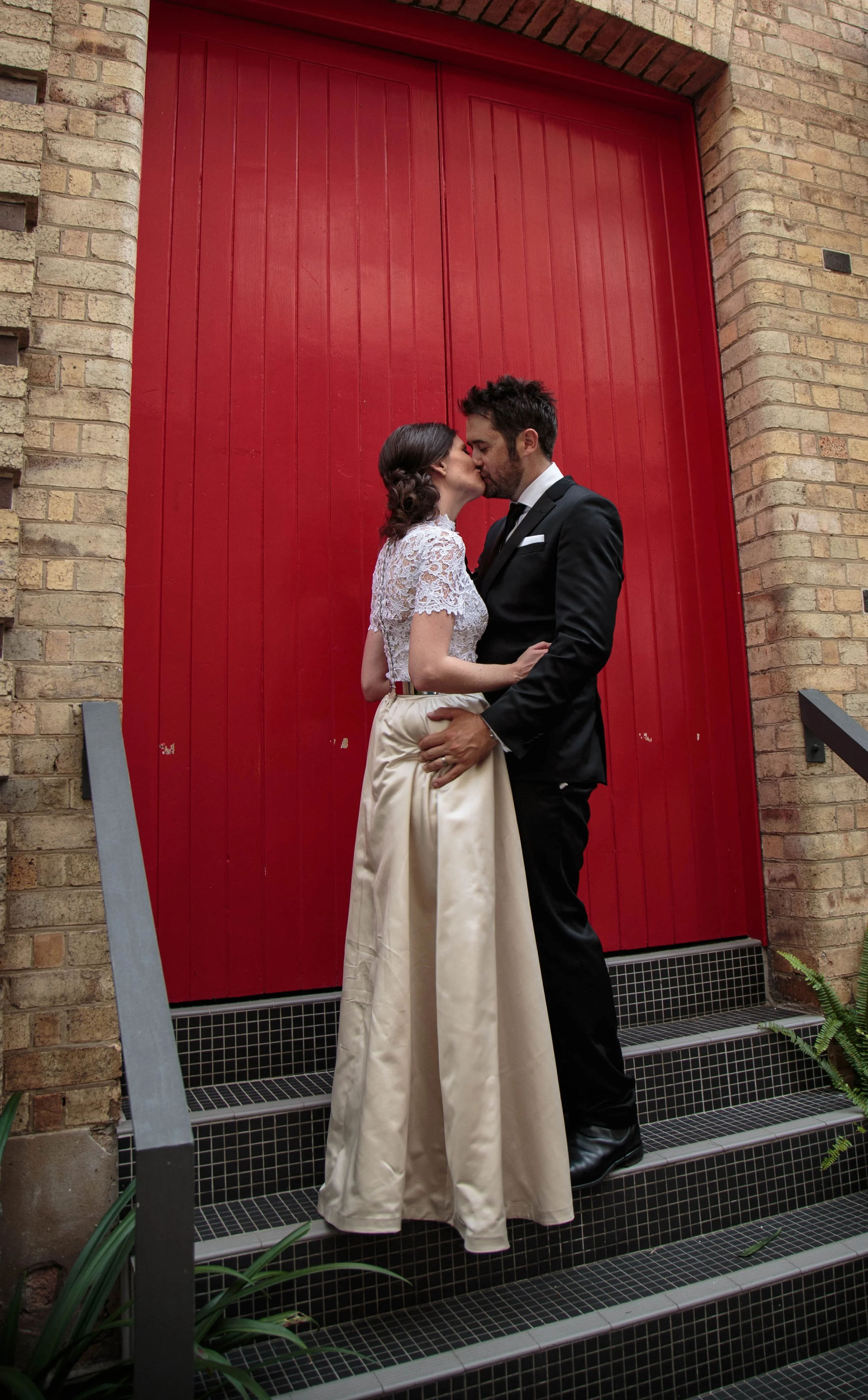 A couple dressed in wedding attire sharing a kiss on a staircase in front of a red door.