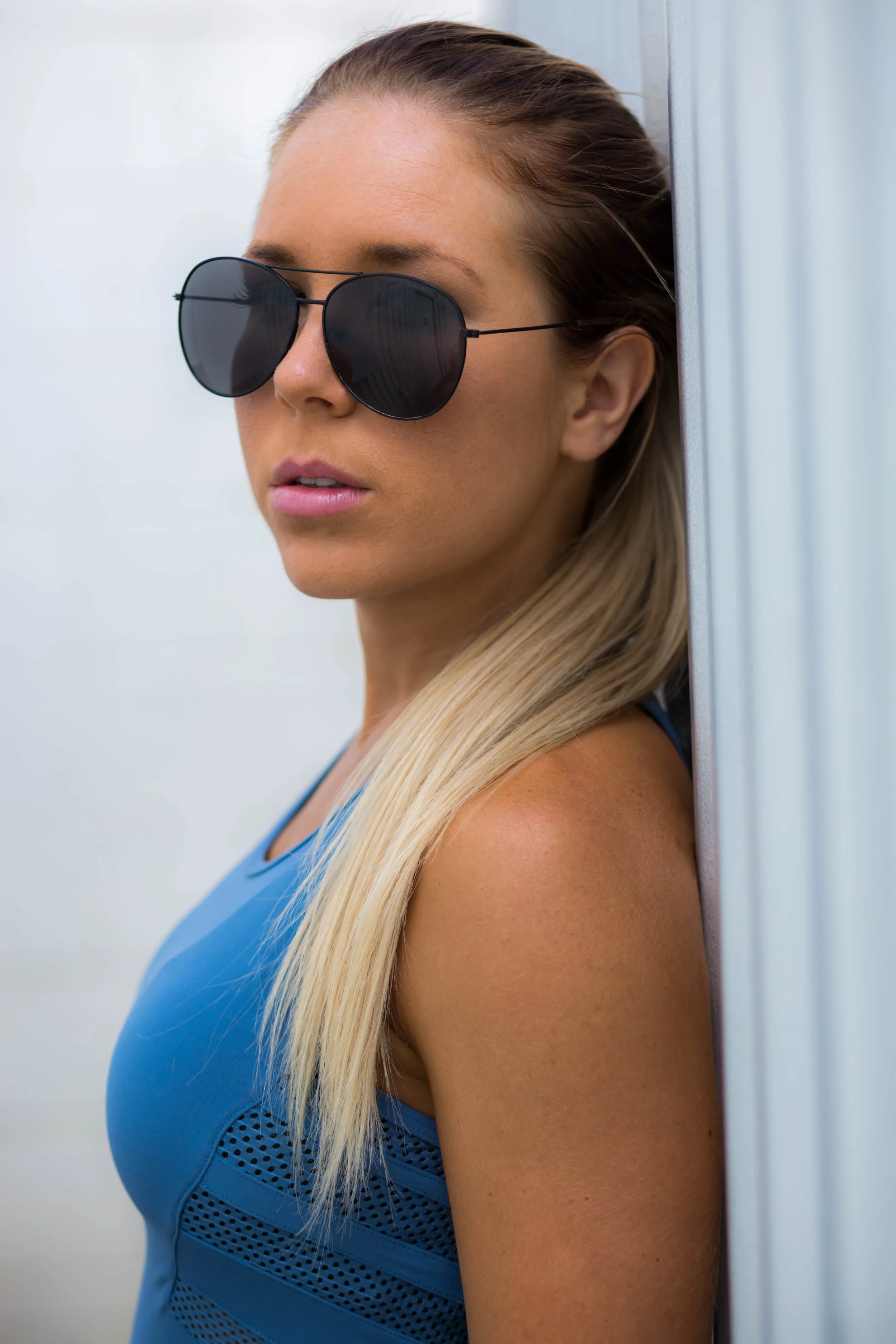 A woman with blonde hair wearing black sunglasses and a blue sports top leaning against a wall.
