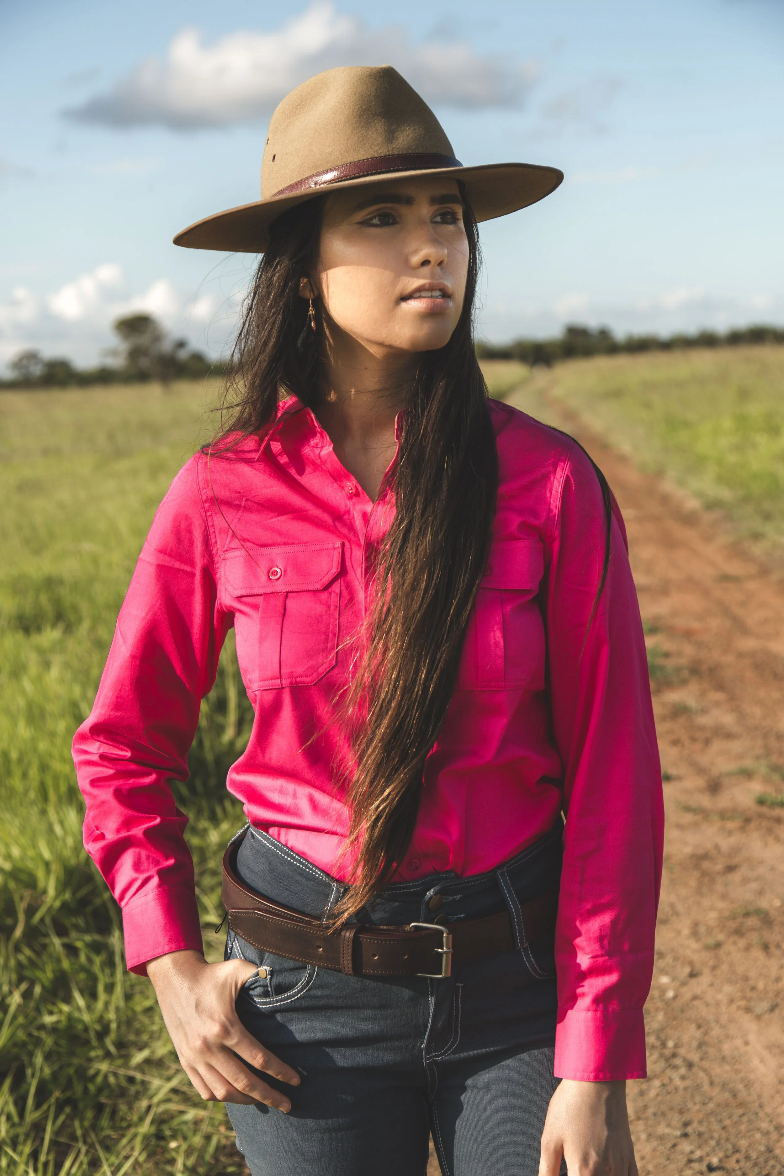 Woman wearing a brown hat, pink shirt, and dark jeans standing on a dirt path in a grassy field under a partly cloudy sky.