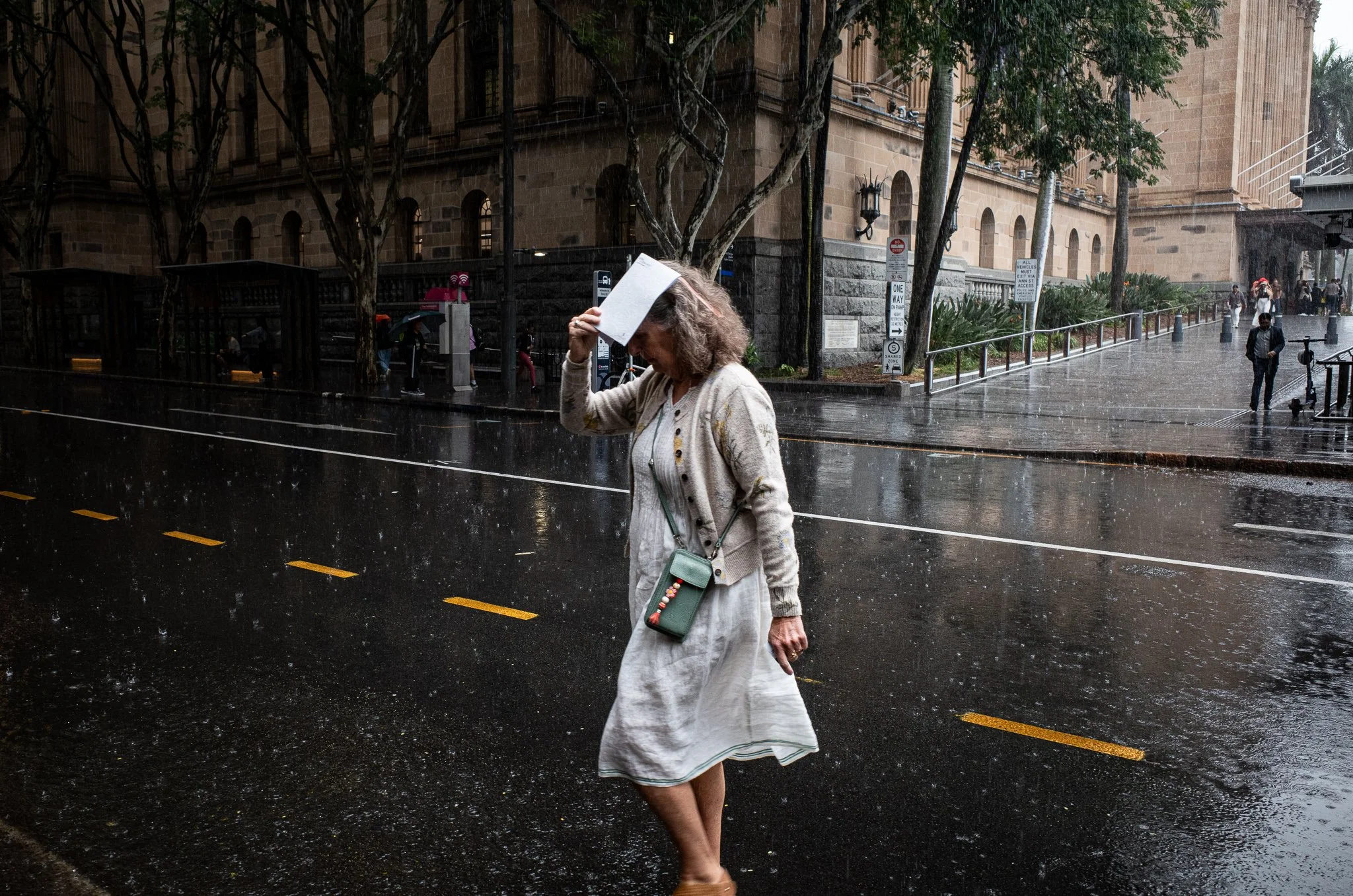 A woman in a white dress and beige cardigan walks barefoot on a rainy city street, holding a book to her head. In the background, there are trees, a building, and pedestrians using umbrellas.