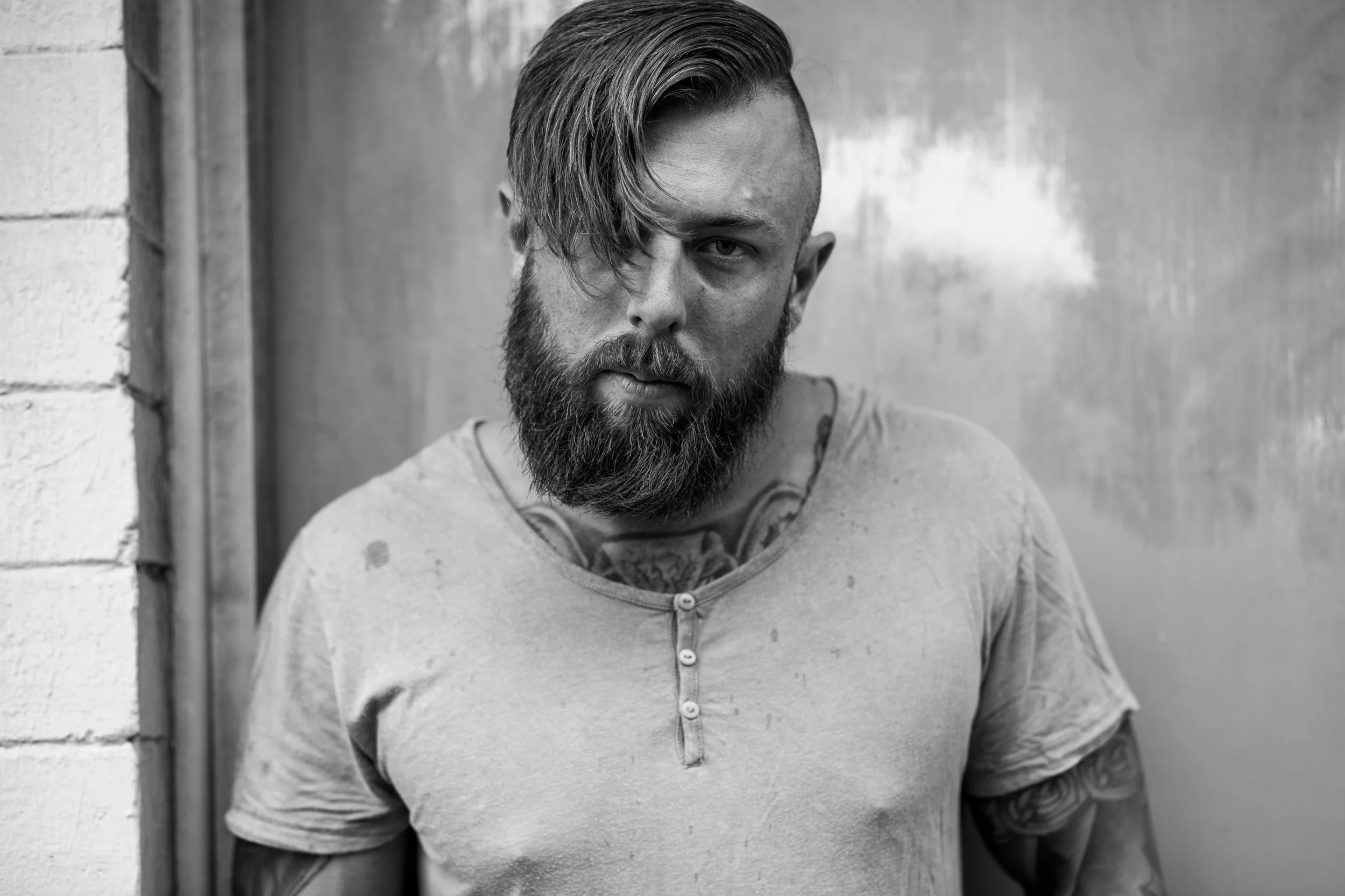 Black and white photo of a man with a beard and styled hair, looking directly at the camera, wearing a casual t-shirt with smudges, standing against a wall.
