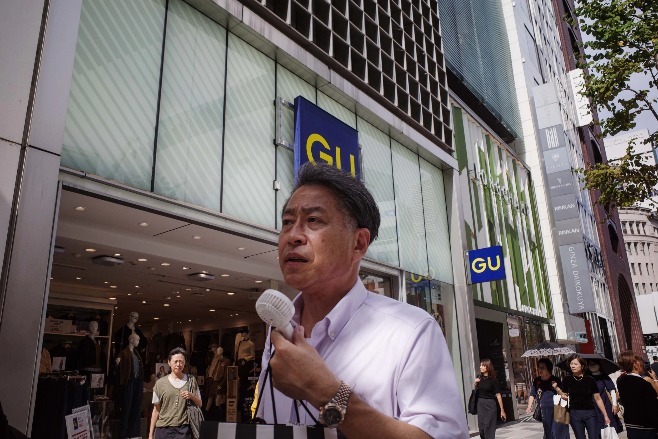 A man in a white shirt holding a small portable fan on a busy street in front of a GU store, with several pedestrians walking by and modern buildings in the background.