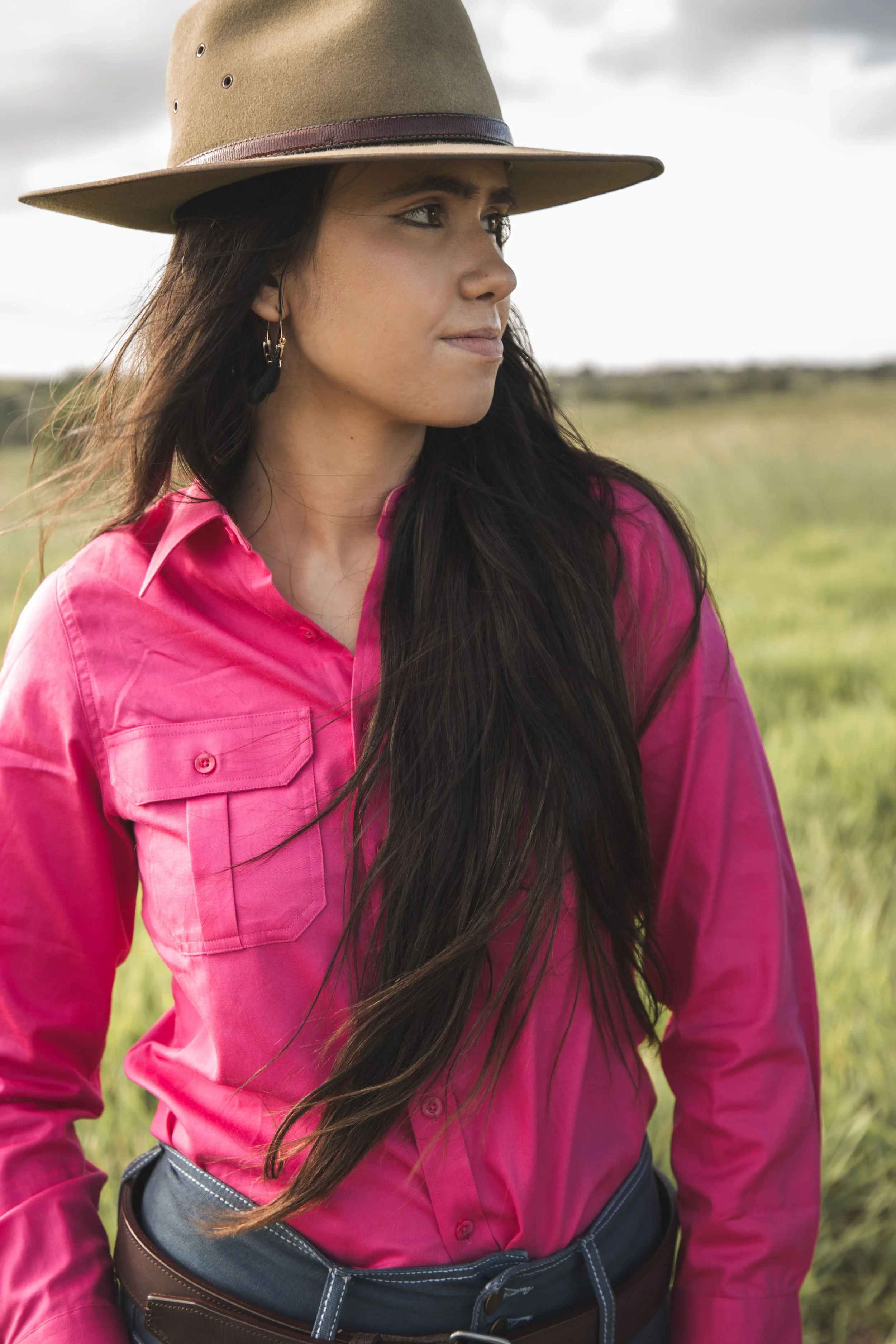A woman in a pink shirt and wide-brimmed hat standing outdoors in a grassy field.