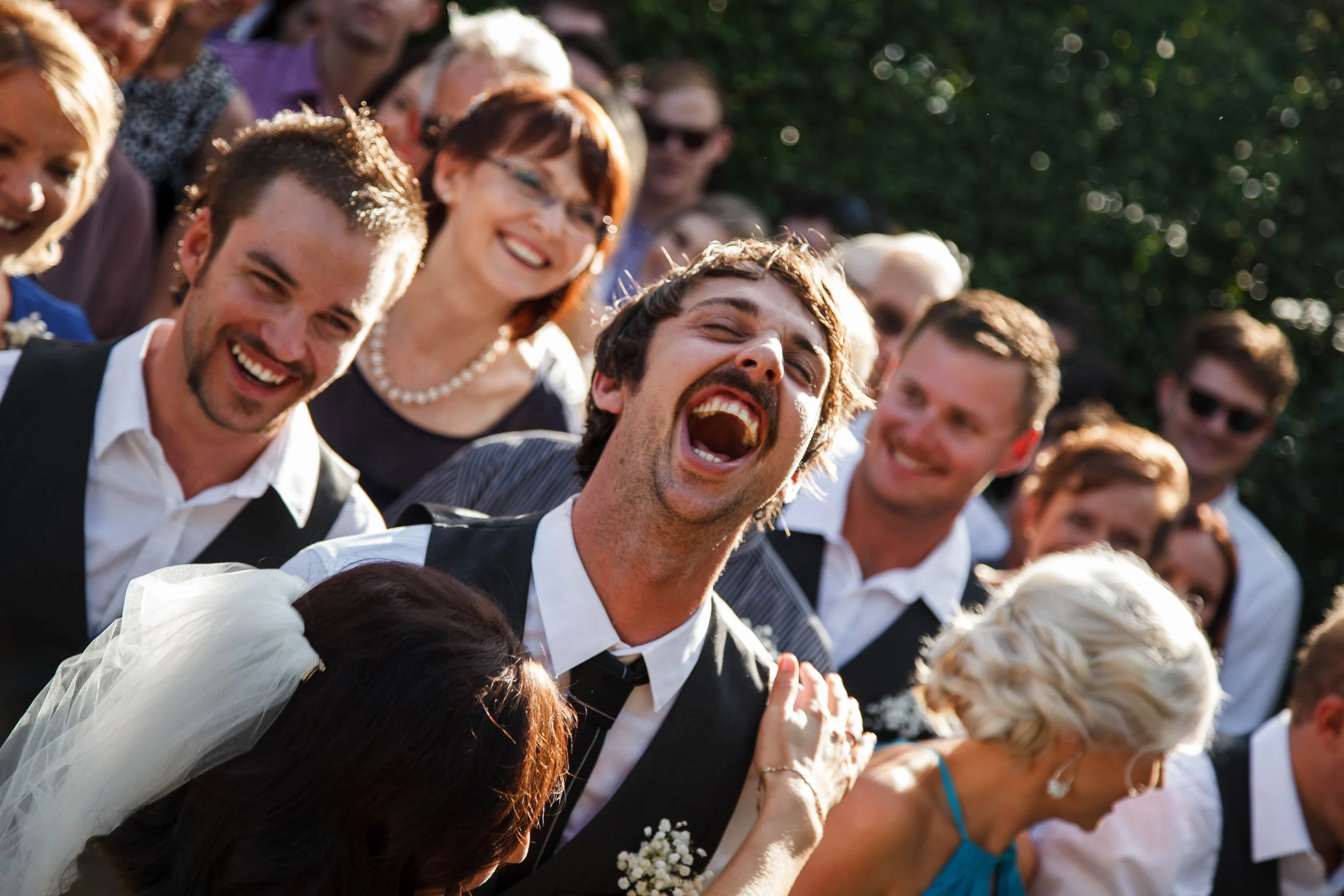 Group of smiling wedding guests outdoors, with the groom laughing, wearing formal attire and surrounded by friends and family.