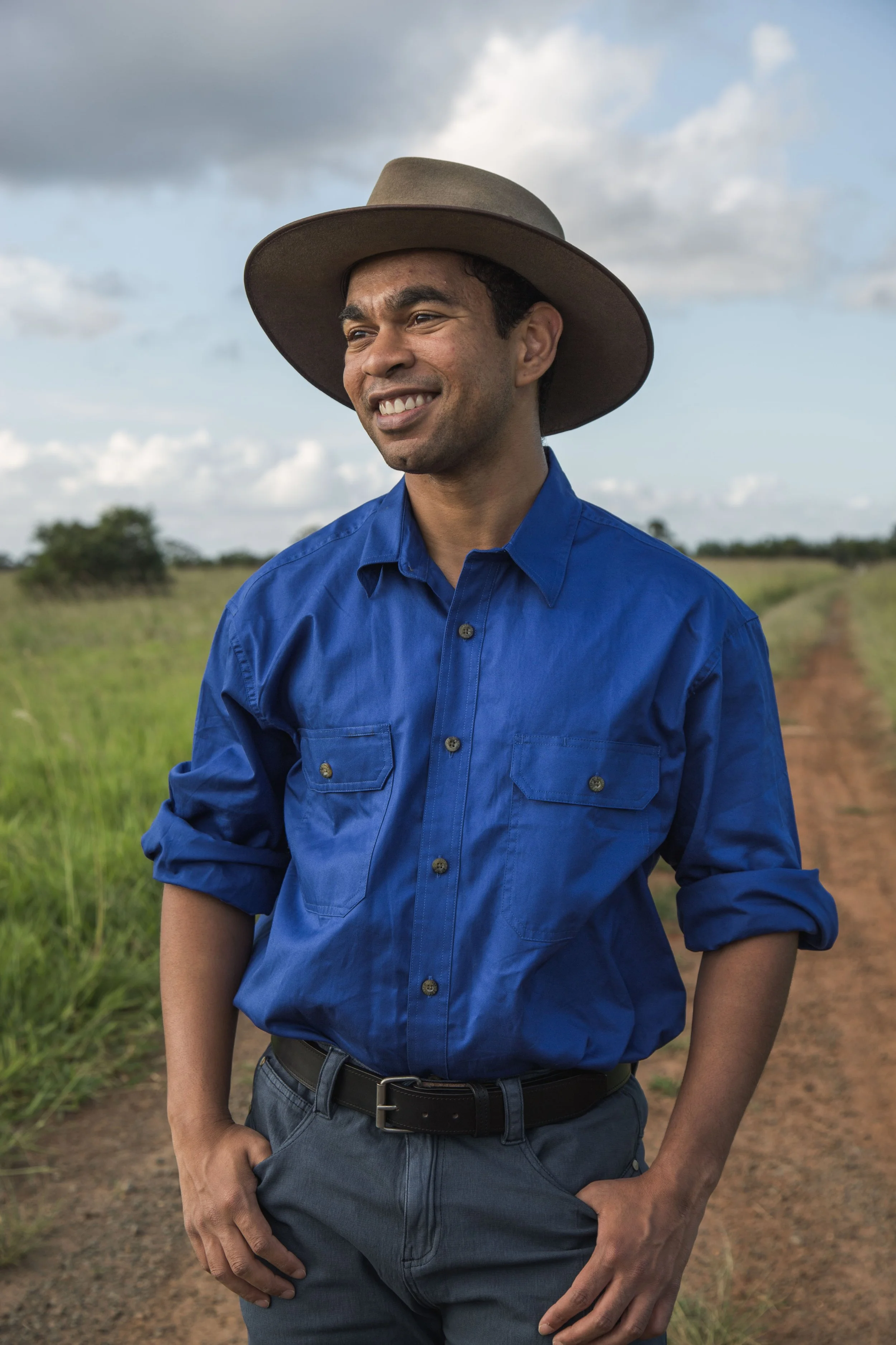 A man wearing a blue shirt and a wide-brimmed hat standing on a dirt path in a grassy field with a cloudy sky in the background.