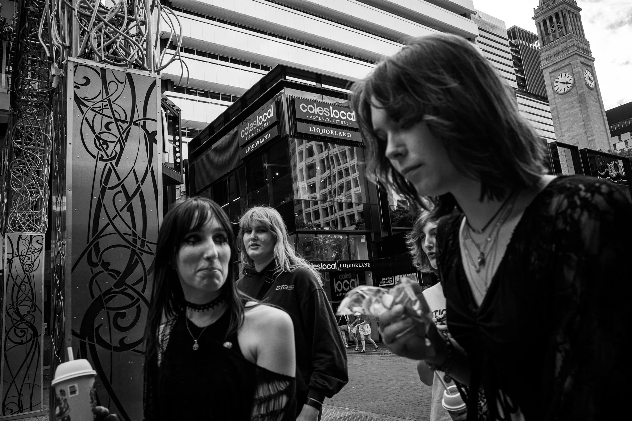 Four young women standing outdoors in a city, with tall modern buildings and a clock tower in the background. The women are dressed in alternative fashion, with one holding a coffee cup and another holding some food, possibly ice cream.
