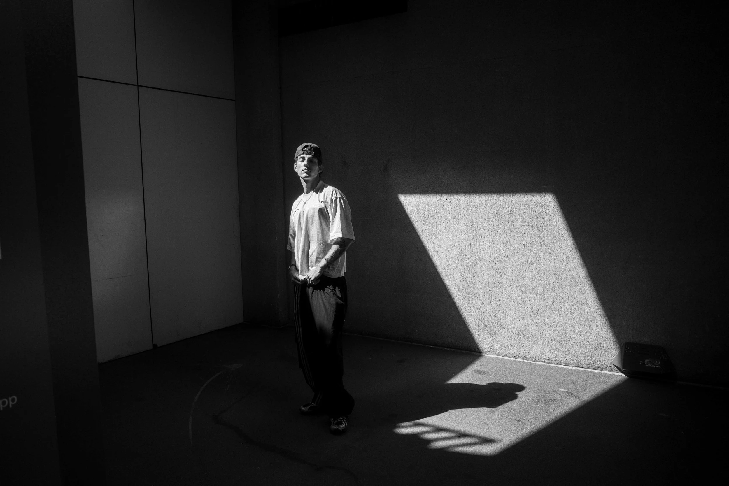 A man stands in a dimly lit room with a large shadow cast by a window on the wall behind him. The image is in black and white.