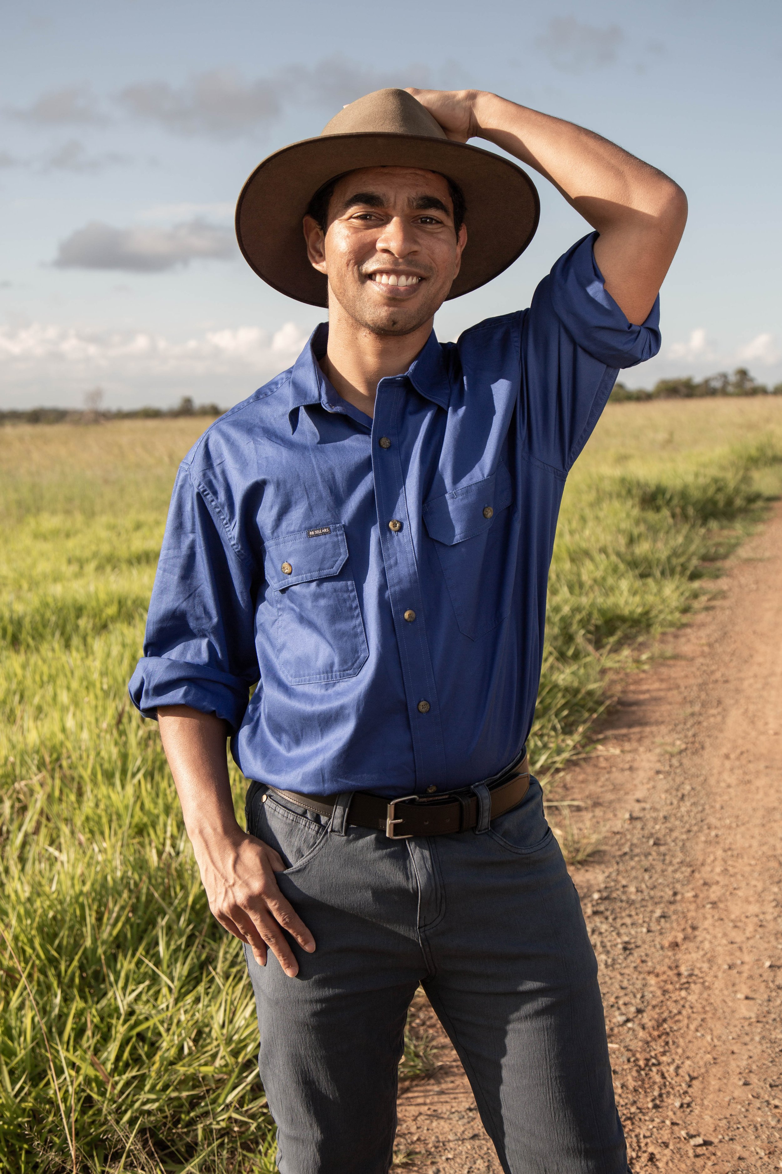 A smiling man in a blue shirt and wide-brimmed hat standing on a dirt path in a grassy field.