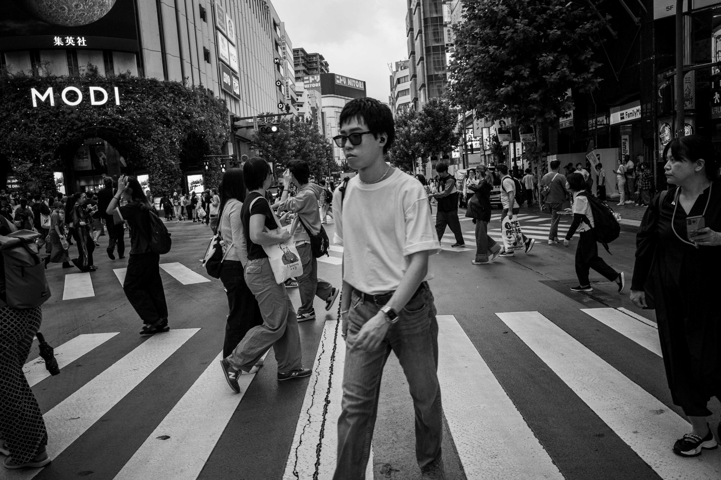 Black and white photo of a busy city crosswalk with numerous pedestrians walking in different directions, tall buildings with storefronts and advertisements in the background, including a visible sign that says 'MODI'.