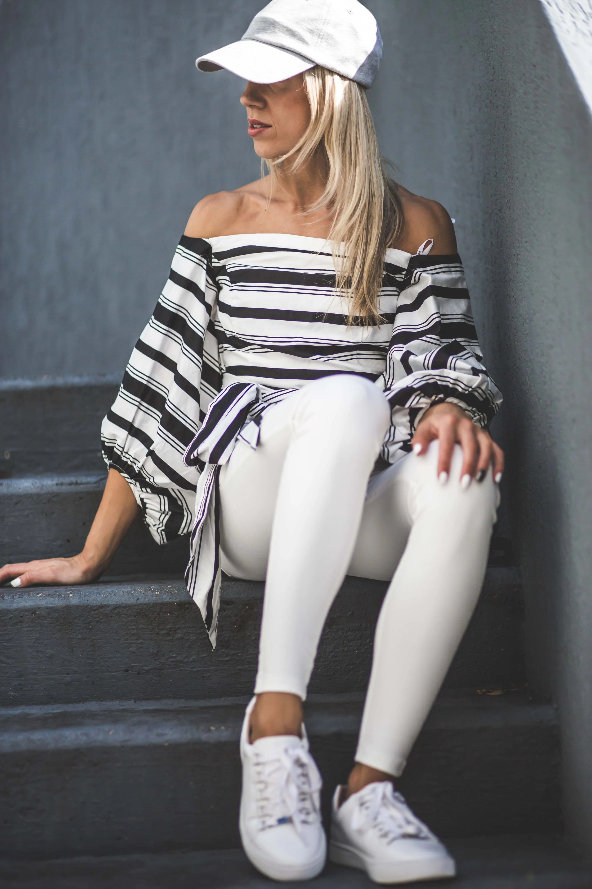 A woman sitting on dark gray stairs, wearing a white baseball cap, off-the-shoulder black and white striped blouse, white leggings, and white sneakers. She has blonde hair and is looking to her left.